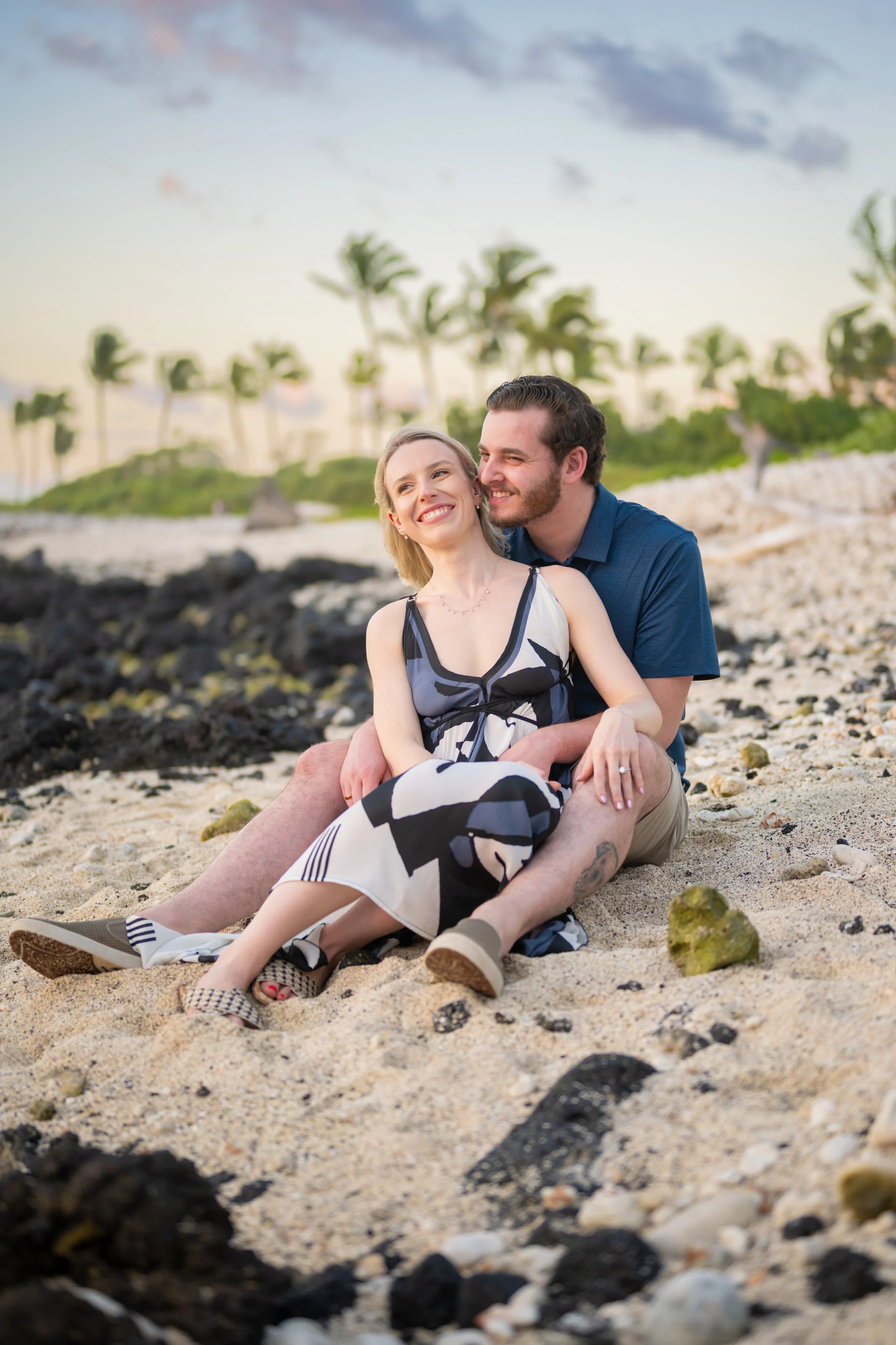 A young couple sitting on a beach with palm trees and rocks in the background during sunset. They are smiling and embracing each other, enjoying a happy moment.