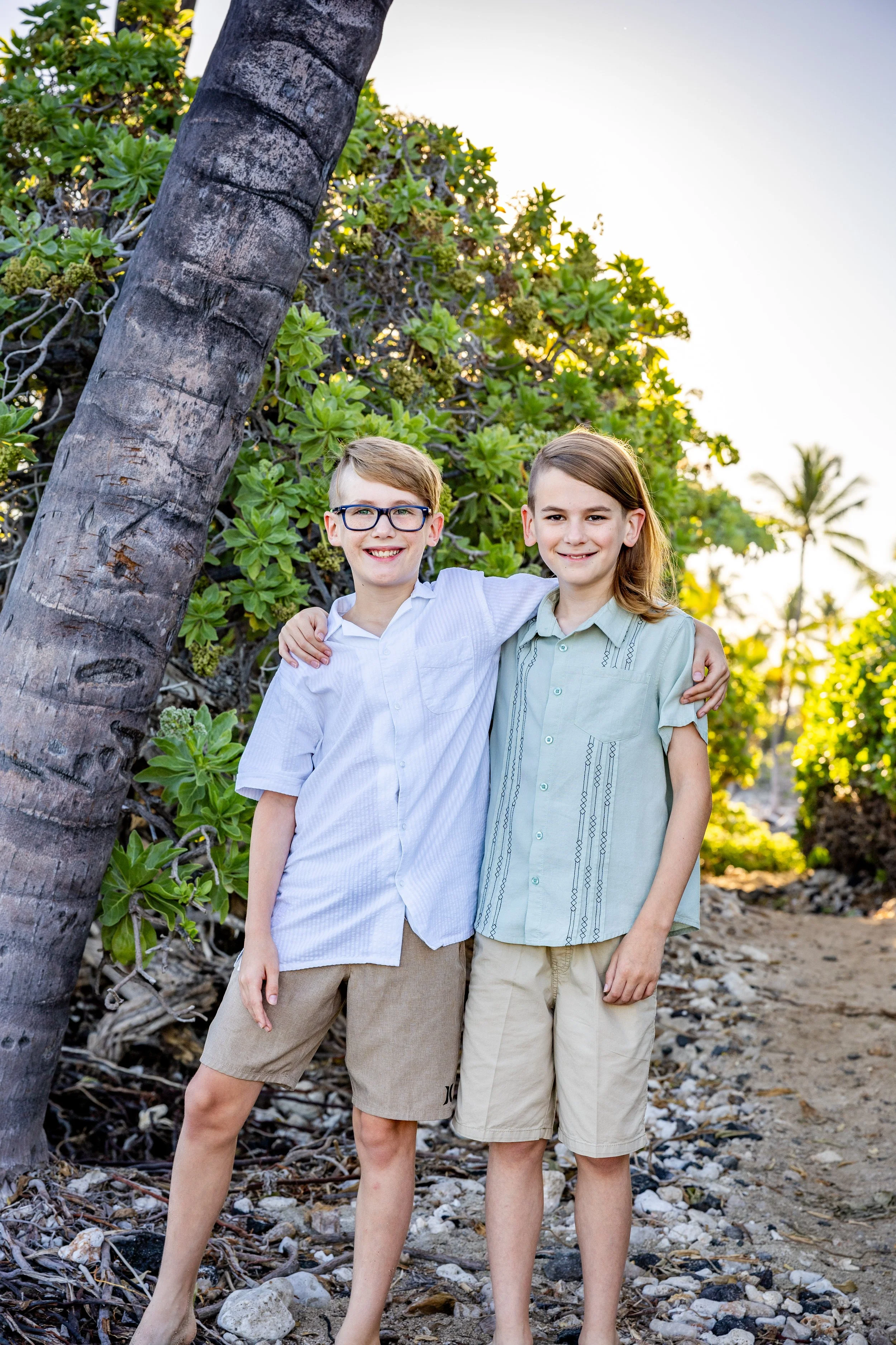 Two young boys standing outdoors on a rocky path, smiling with their arms around each other, near lush green bushes and palm trees, with the sun setting in the background.