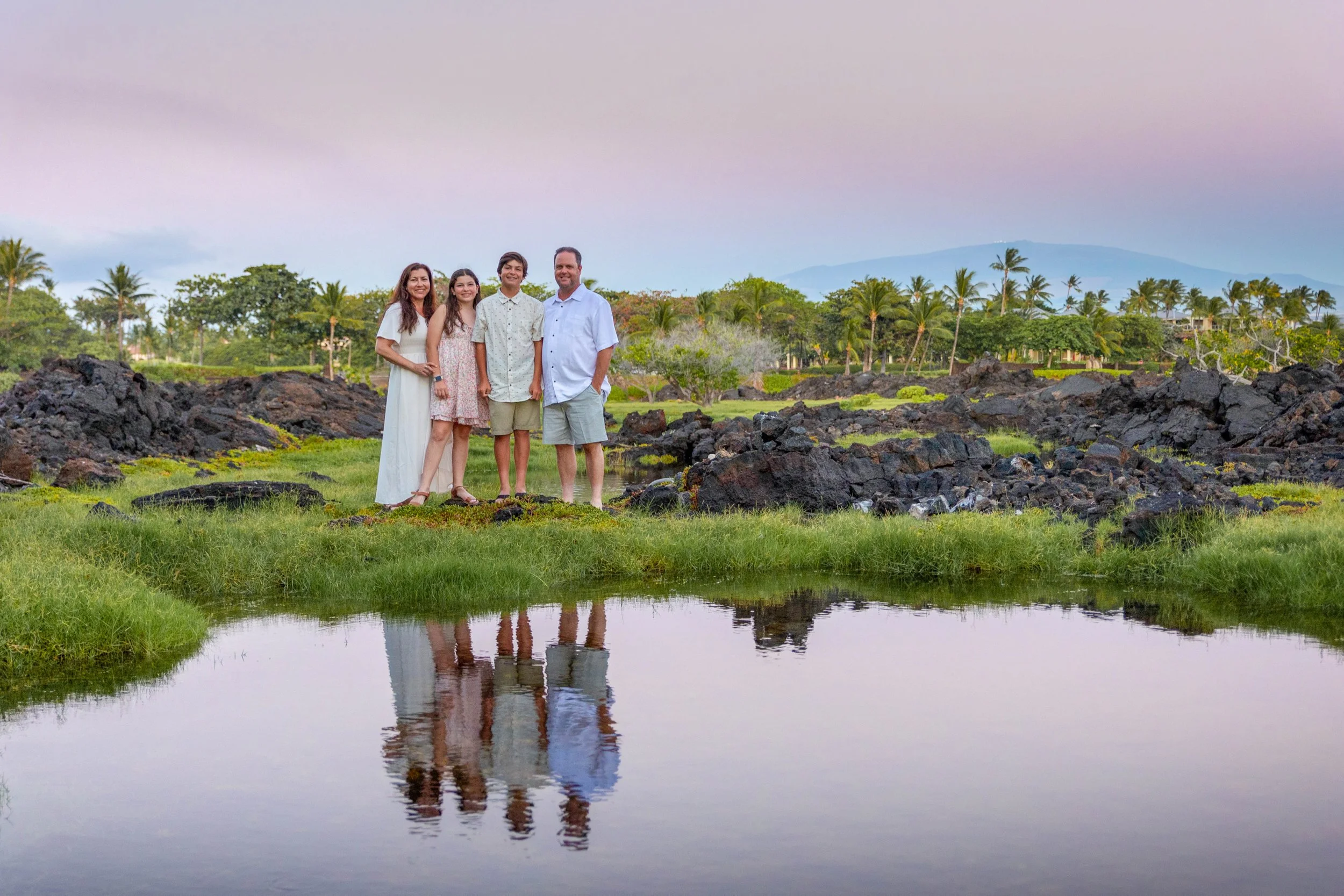 Family of four standing outdoors near a pond with rocky terrain and lush greenery, trees, and mountains in the background.