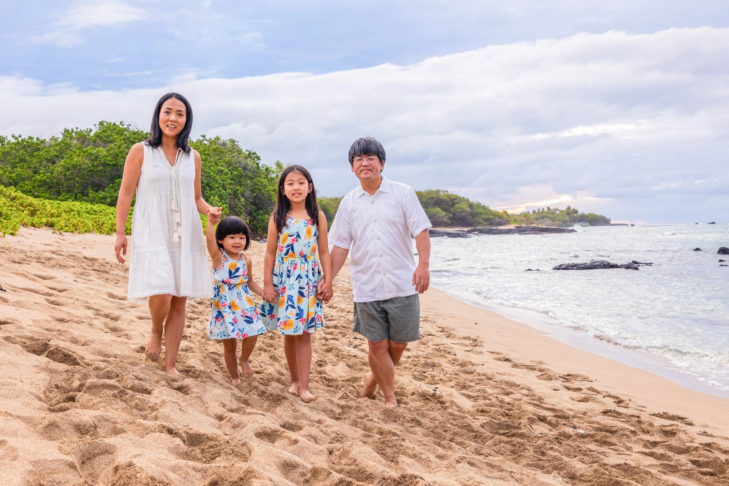 Family of five walking along a sandy beach, holding hands, with ocean waves and cloudy sky in the background.