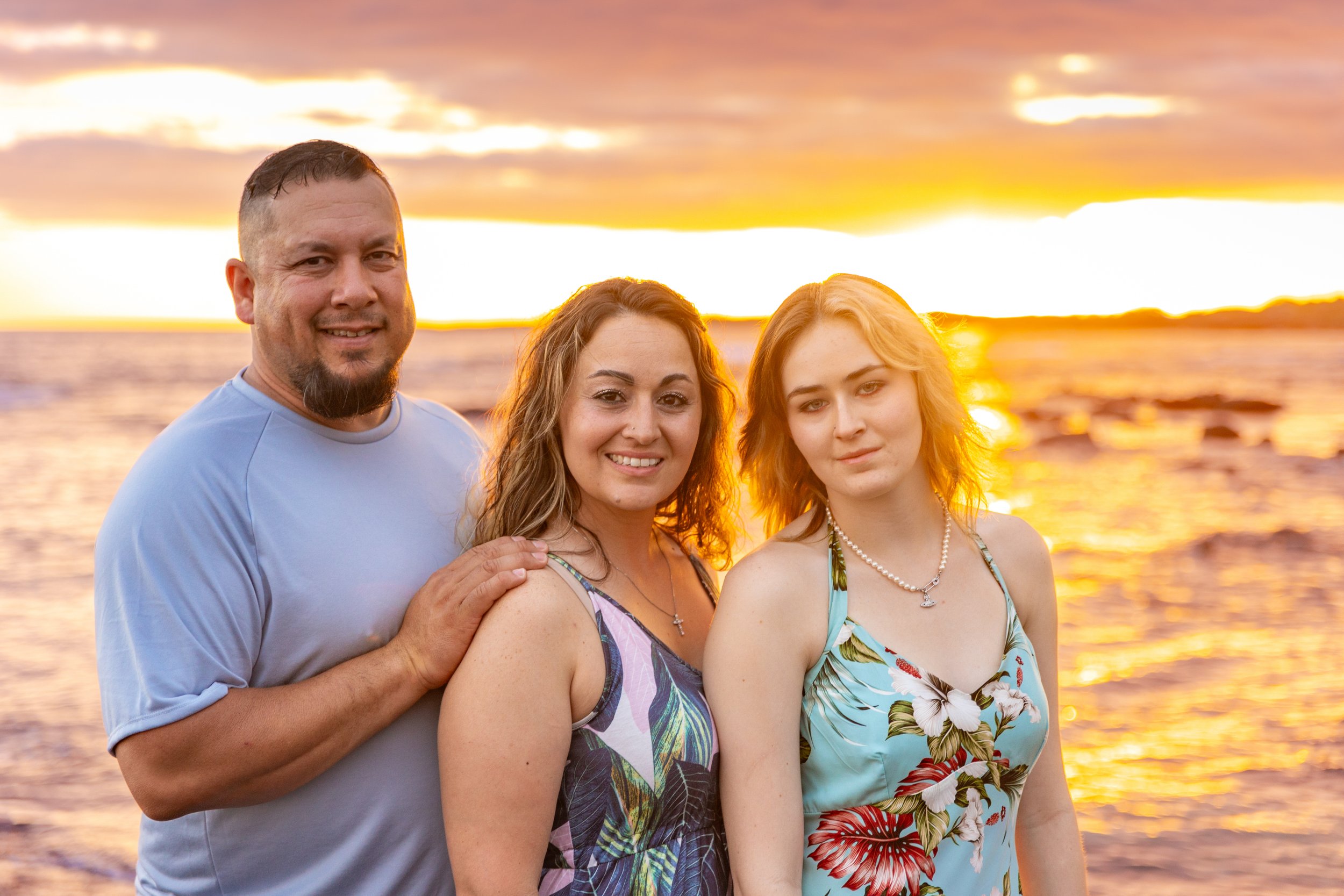 A family of three standing on a beach during sunset, with the ocean and a cloudy sky in the background.