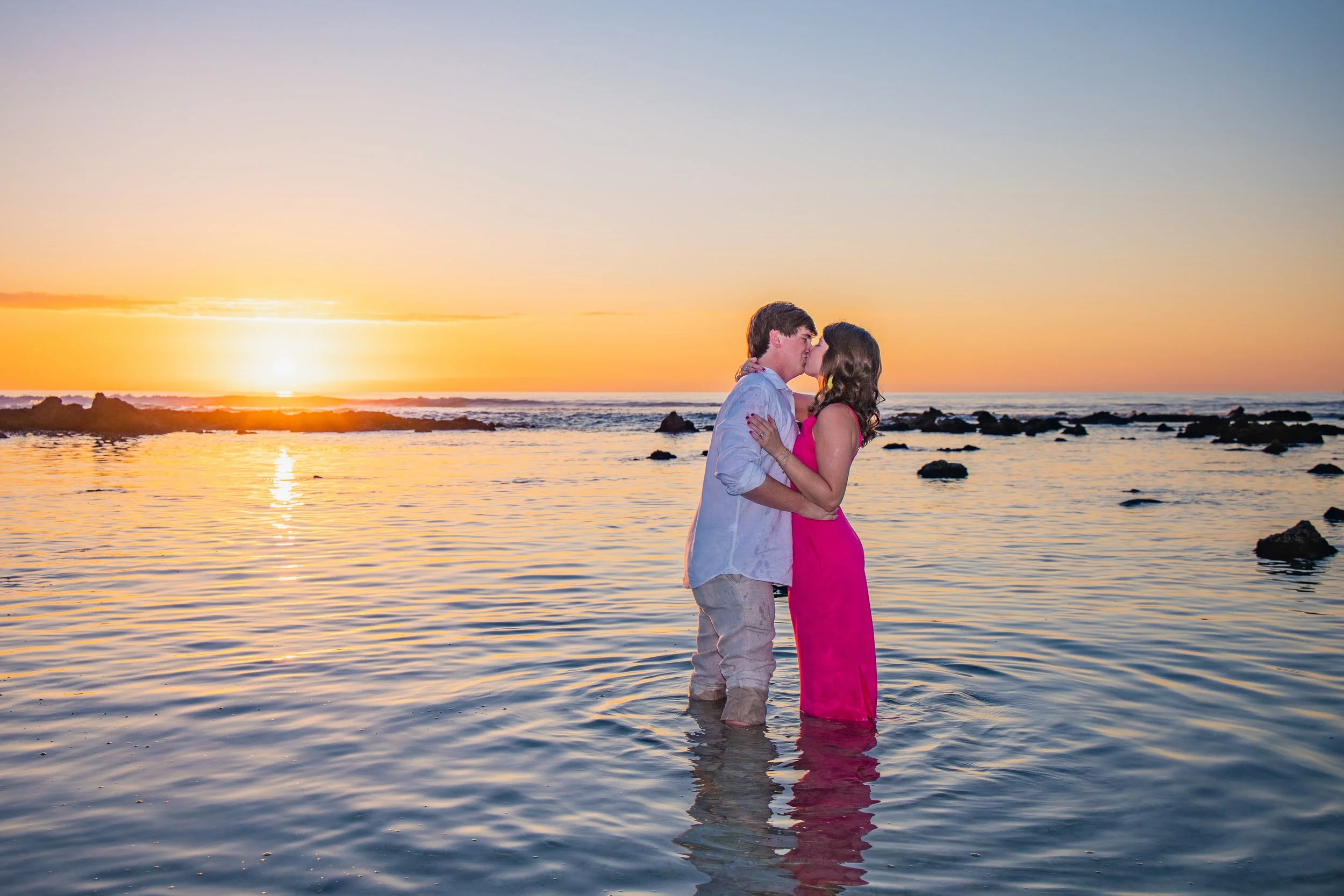 A couple sharing a kiss in the ocean at sunset, with rocks and the setting sun in the background.