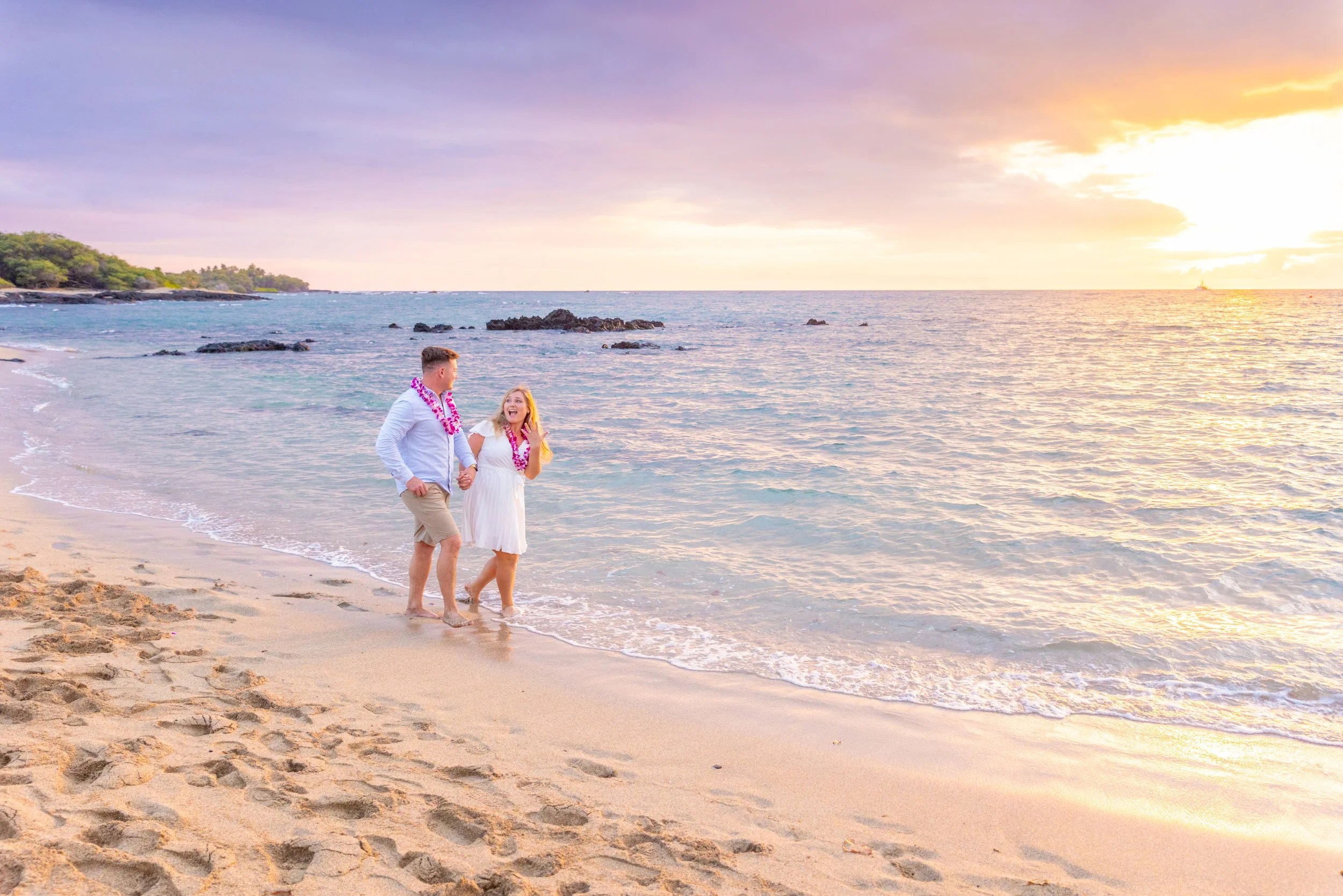 A couple walking hand in hand along a sandy beach at sunset, wearing leis and smiling, with the ocean and sky in the background.