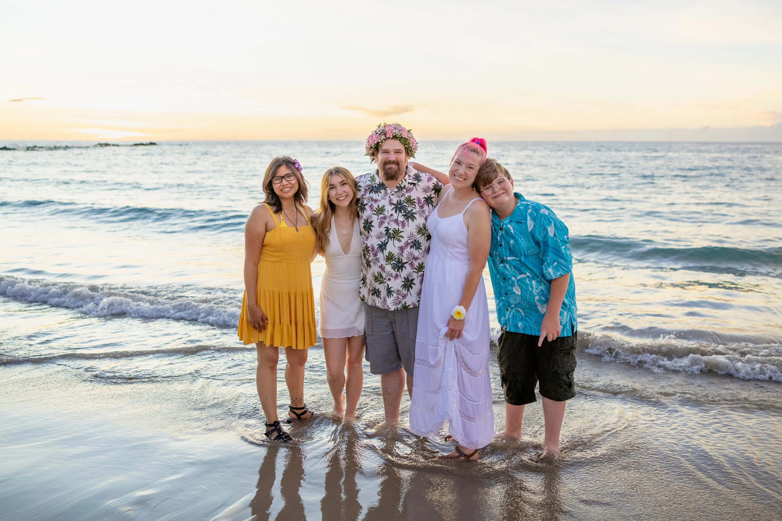 Group of five friends enjoying a sunset on the beach, standing in the shallow water, smiling, with two women wearing white dresses, one woman in a yellow dress, a man with a floral shirt and a flower crown, and a young man in a blue shirt leaning on 