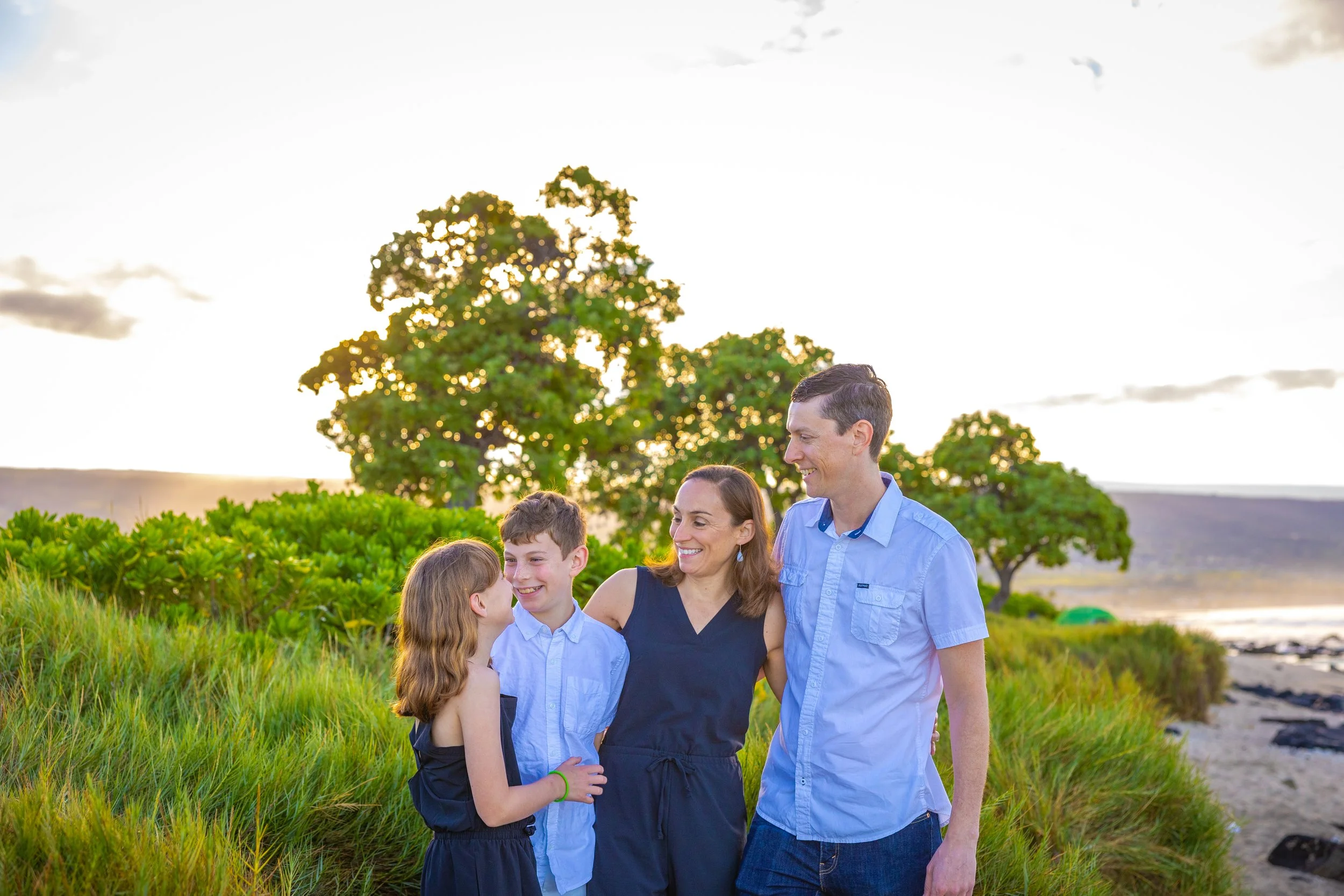 A family of four, including a mother, father, son, and daughter, enjoying time together outdoors on a grassy area near the beach with green trees and a sunset in the background.