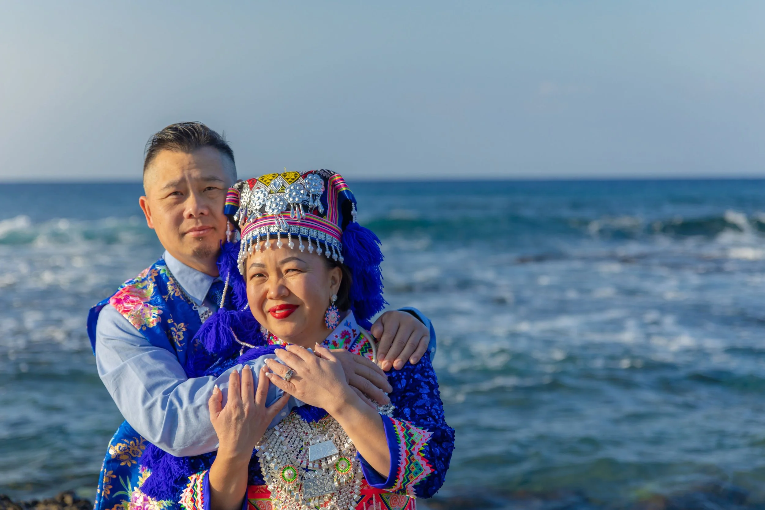 A couple standing on the beach in traditional colorful indigenous clothing, with the ocean in the background.