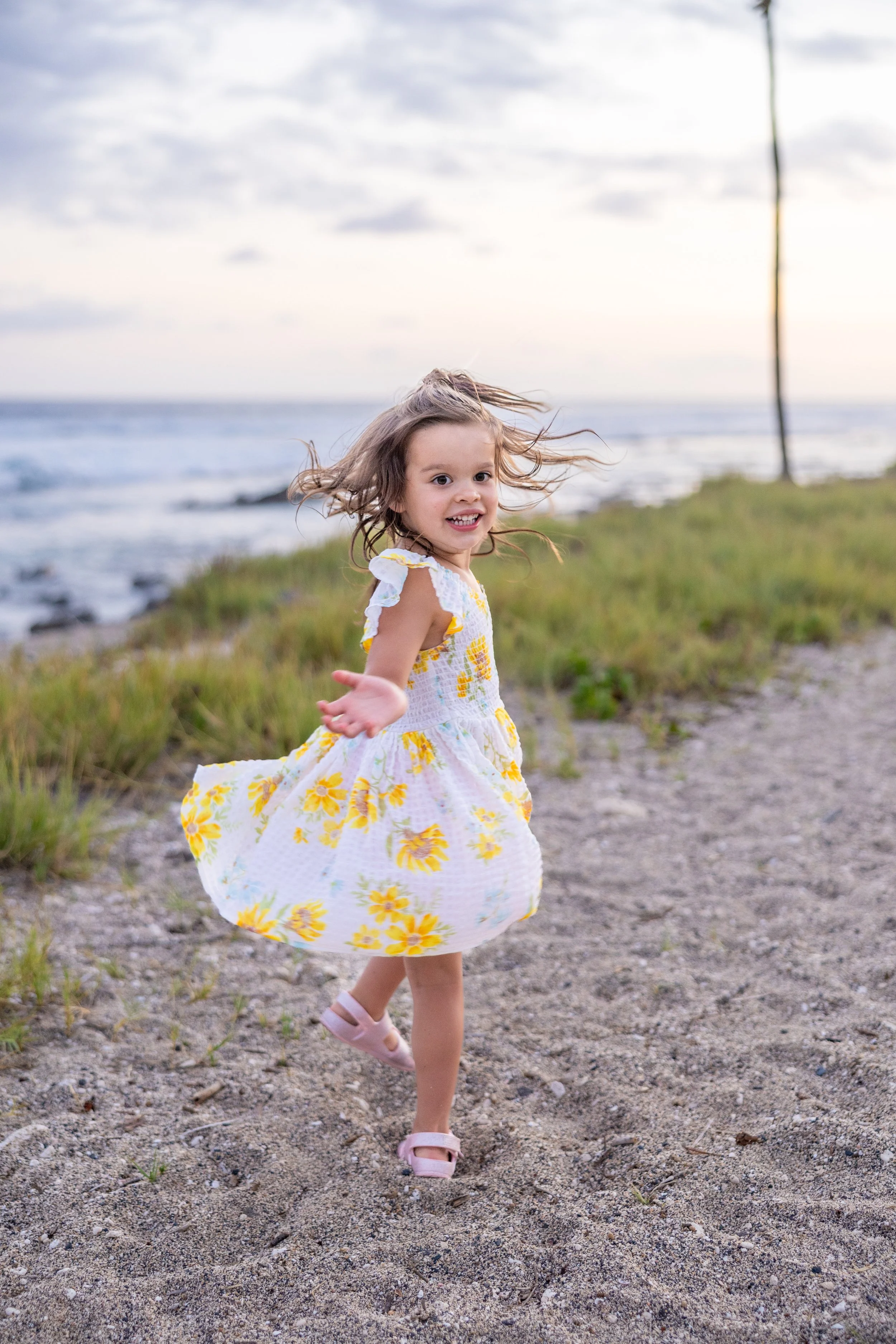A young girl running happily on a beach path during sunset, wearing a white dress with yellow flowers and pink sandals, with her hair flowing in the wind.