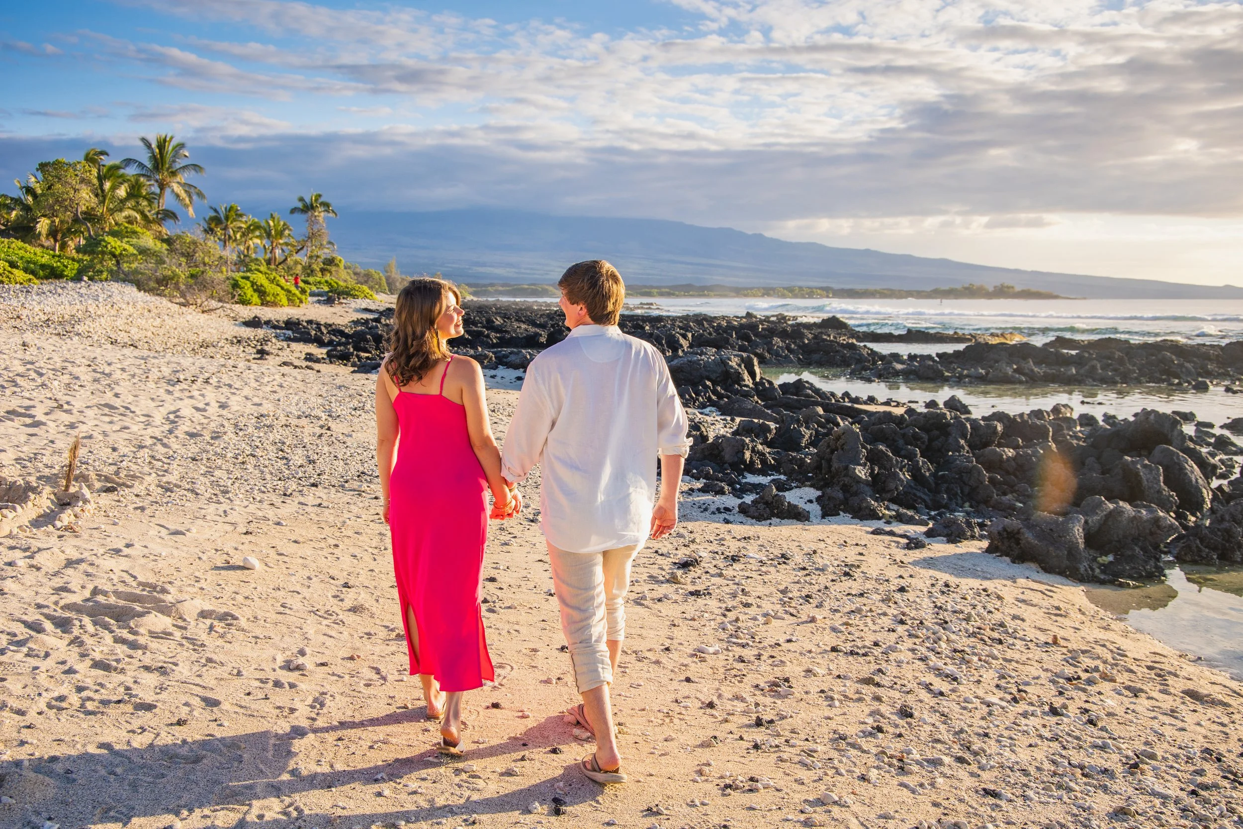 A couple walking hand in hand on a sandy beach near the ocean during sunset, with tropical trees and mountains in the background.