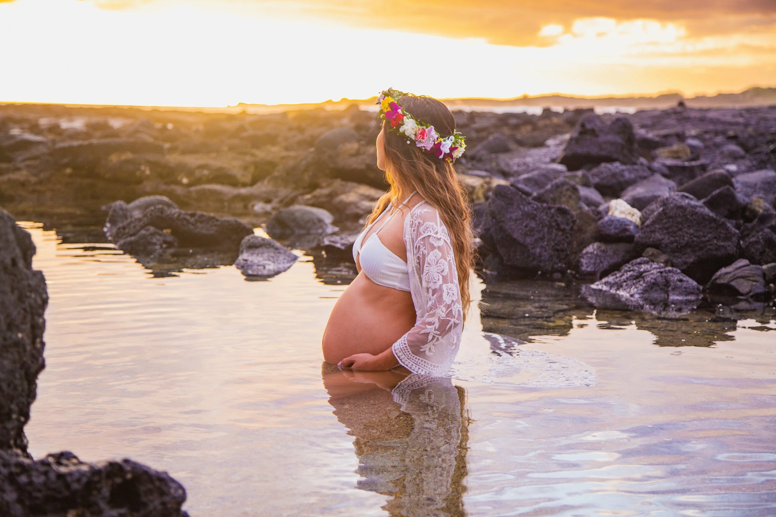 A pregnant woman in a white dress and lace top with a floral crown stands in a body of water during sunset, surrounded by rocks.