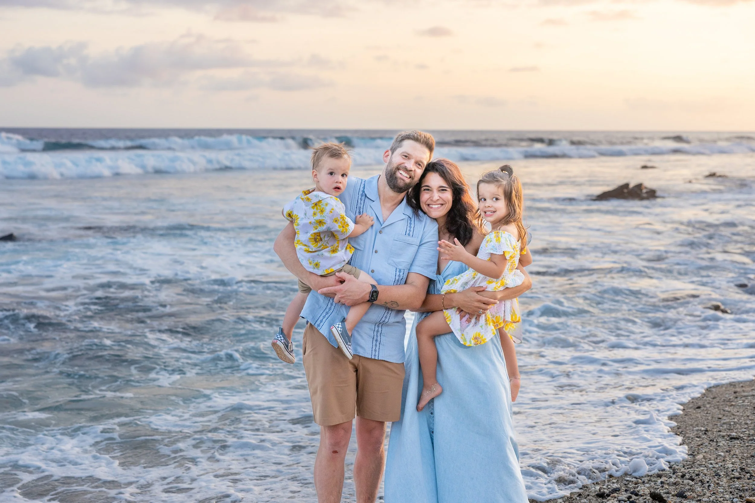 Family of four enjoying time at the beach during sunset. A man, woman, and two young girls are smiling and holding each other near the shoreline with waves and a cloudy sky in the background.