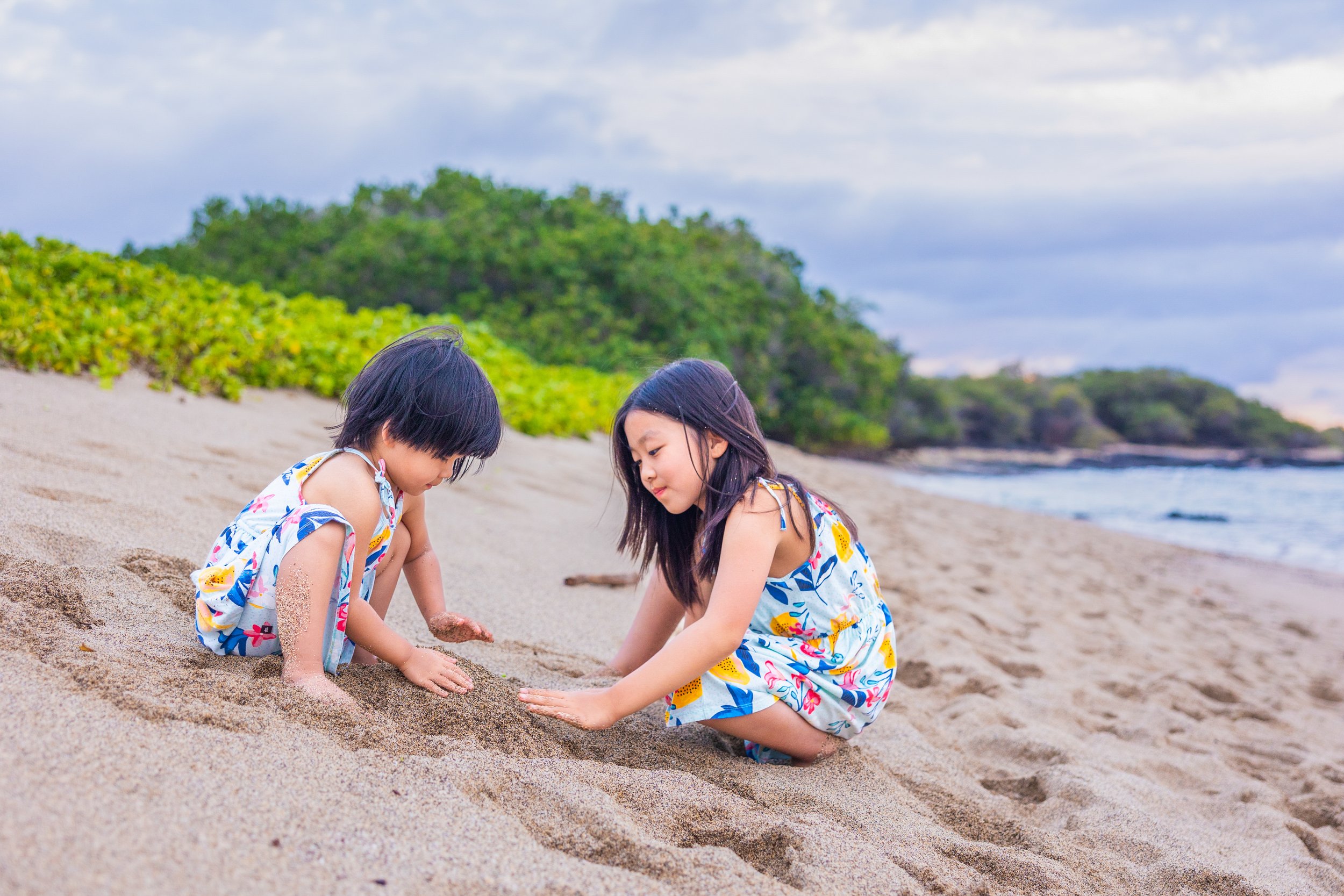 Two young girls in colorful dresses playing in the sand on a beach with green bushes and trees in the background.