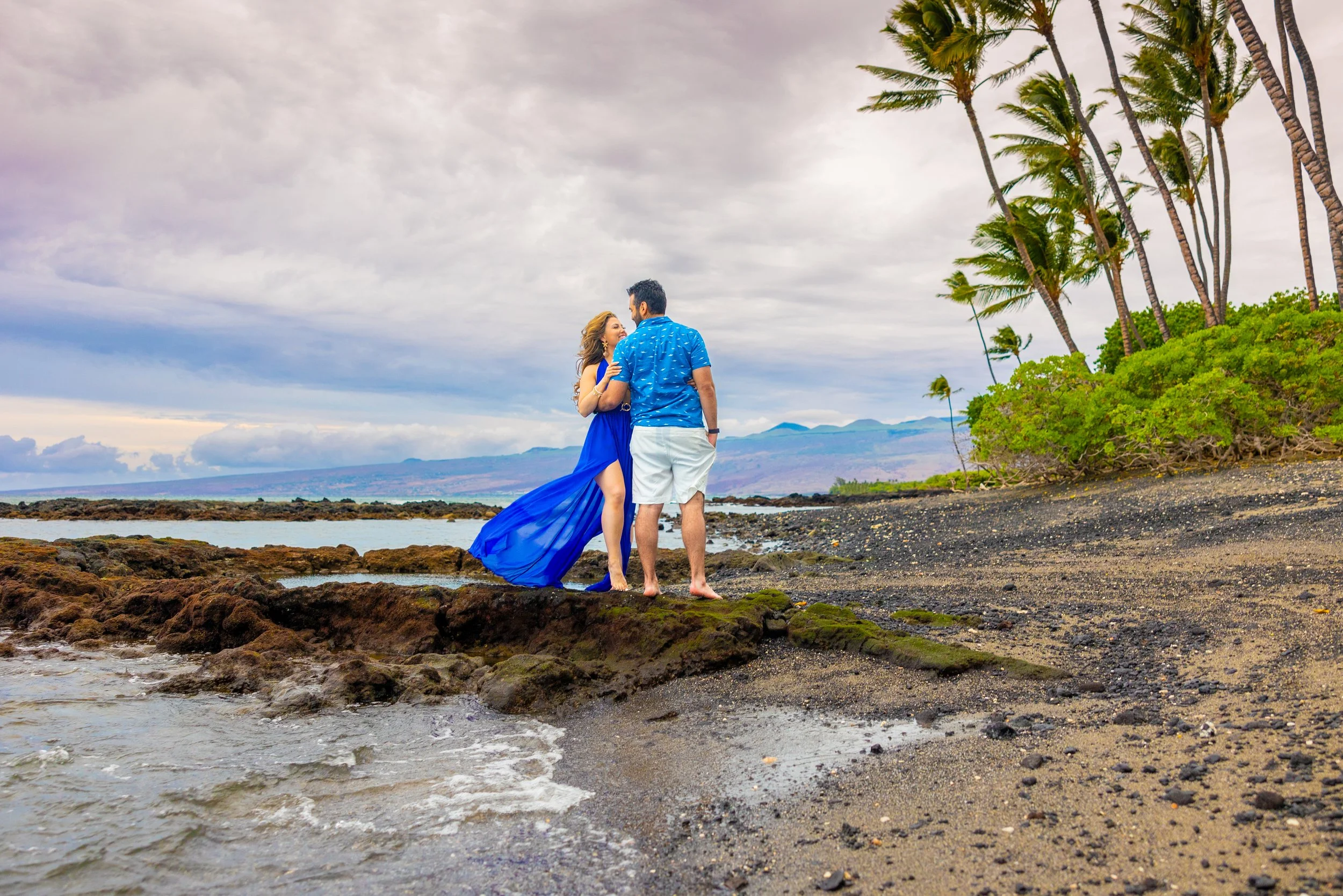 A couple stands on a rocky shoreline, embracing and gazing at each other. The woman wears a flowing blue dress with a slit, and the man wears a blue tropical shirt with white shorts. There are palm trees on the right, and the ocean and mountains are 