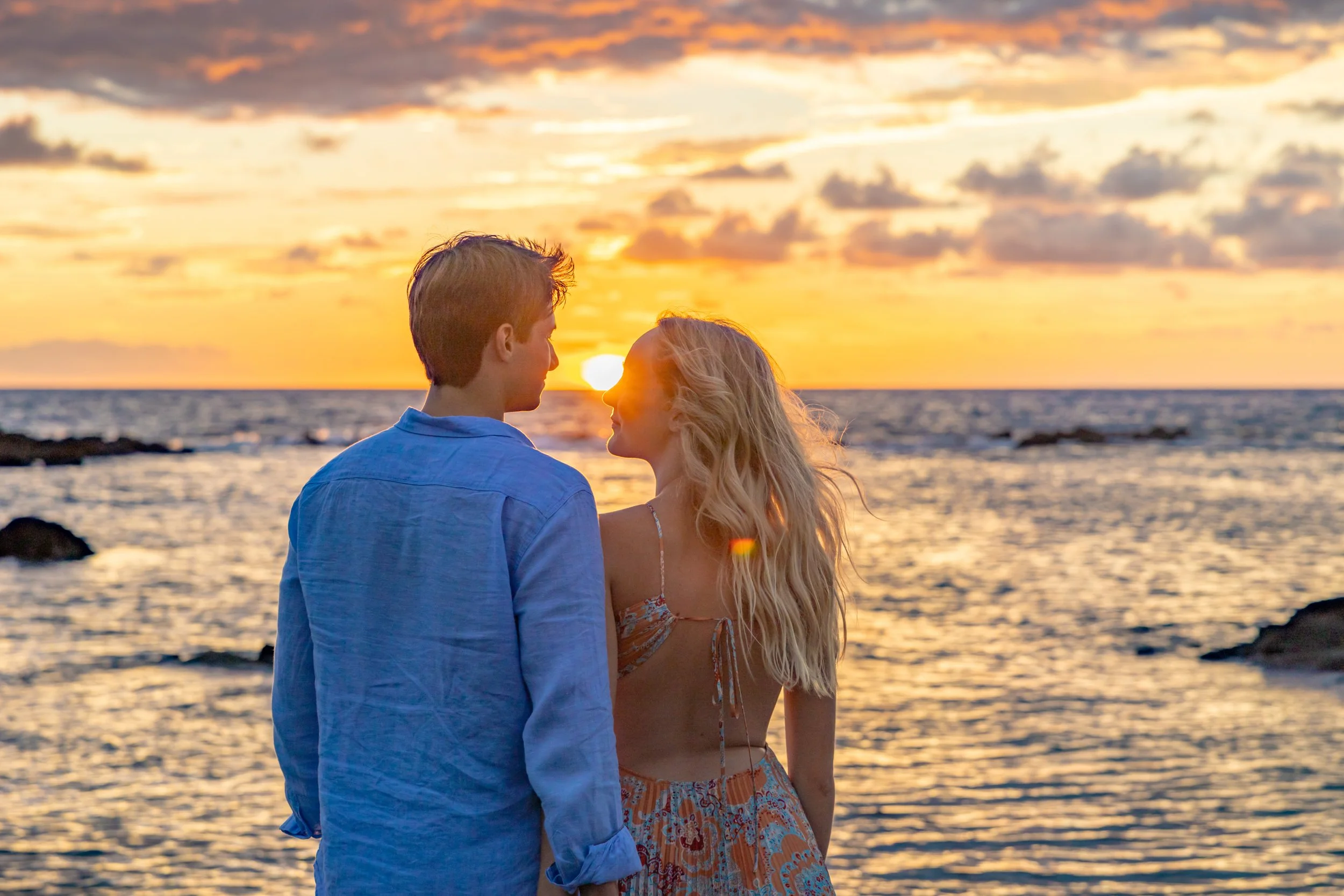 A couple standing on the beach at sunset, facing each other with the sun setting over the ocean in the background.