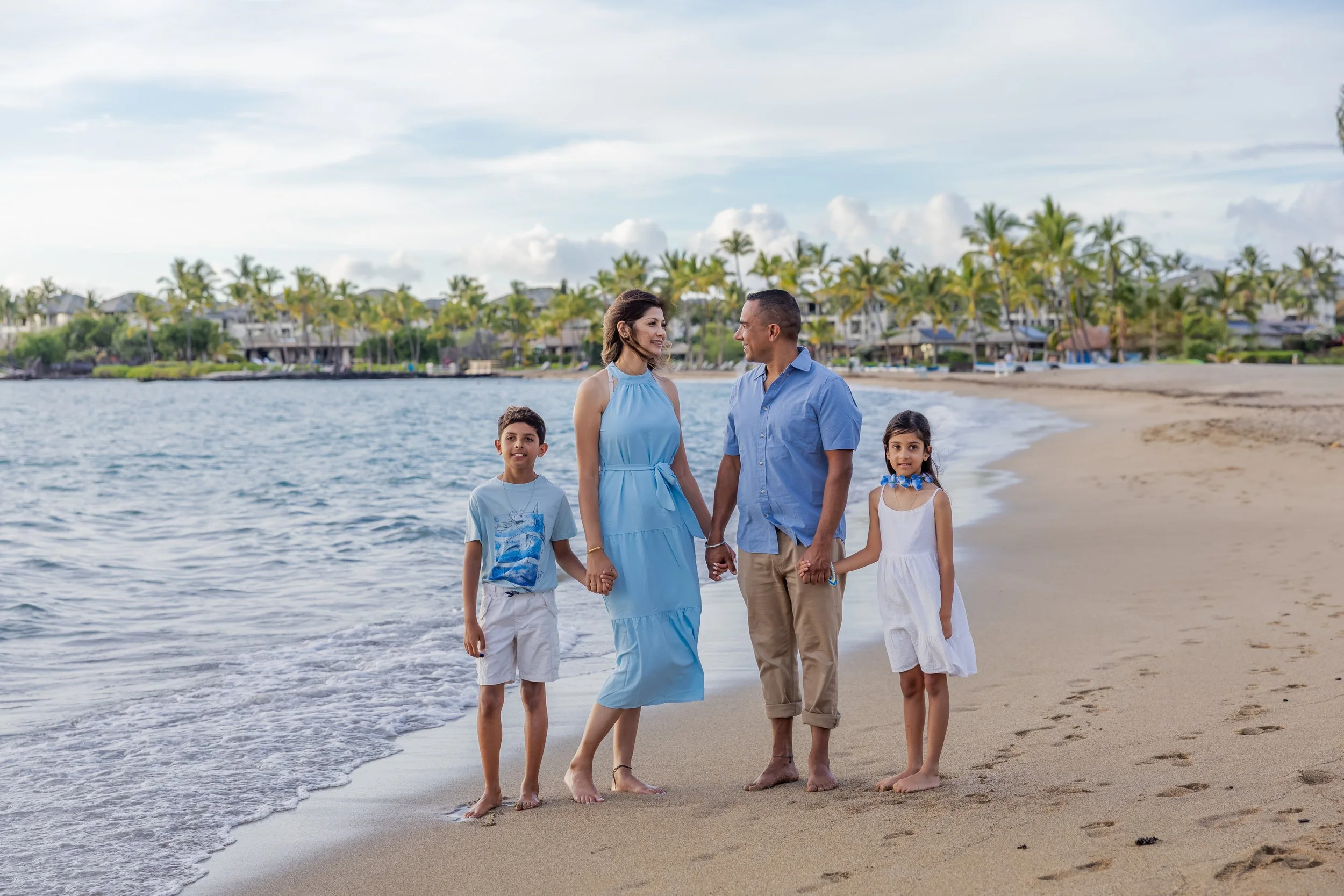 A family of four holding hands walking on a sandy beach near the water, with palm trees and houses in the background.