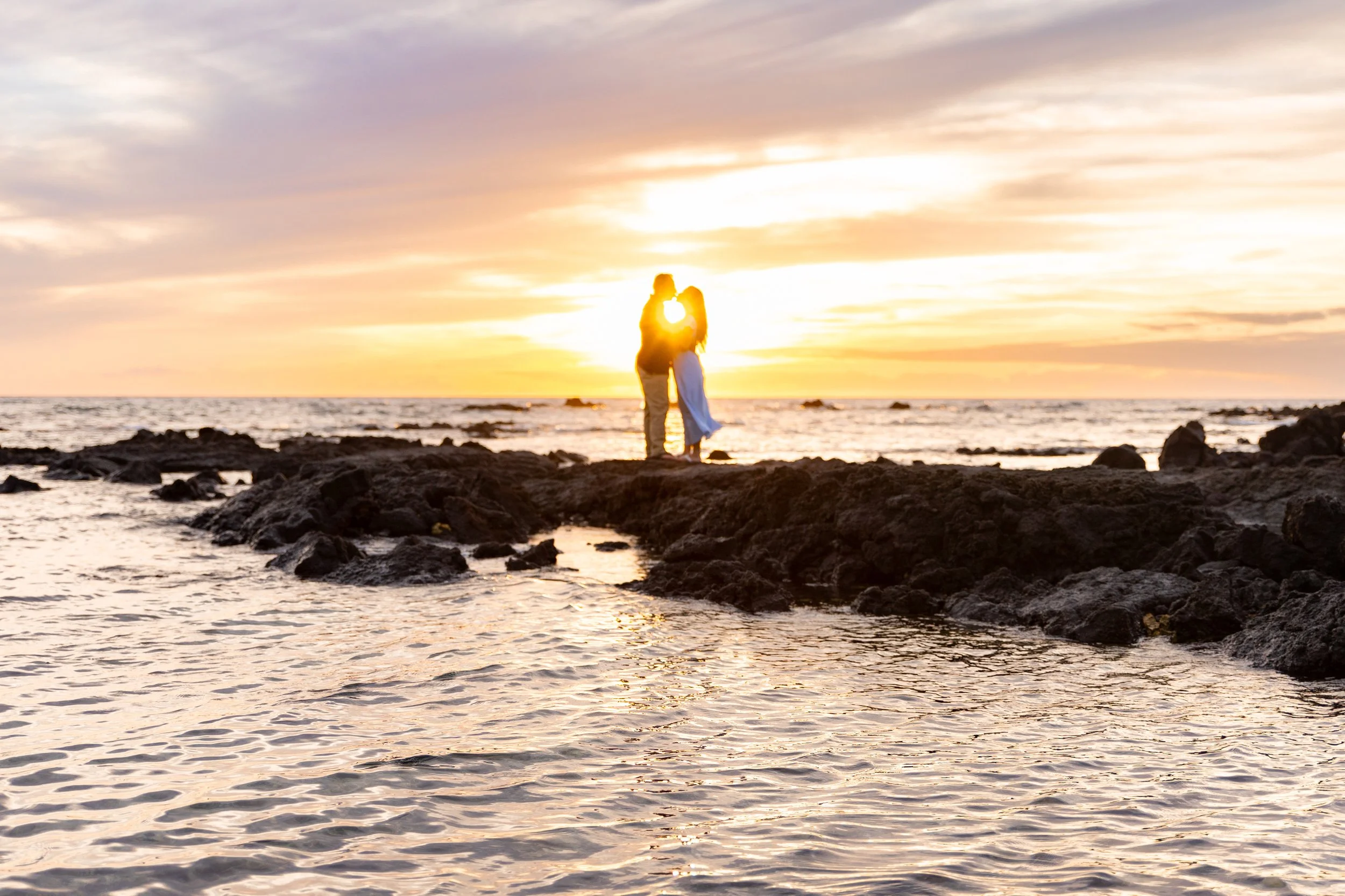 Silhouette of a couple standing on rocks at the beach during sunset, with the sun setting over the ocean behind them.