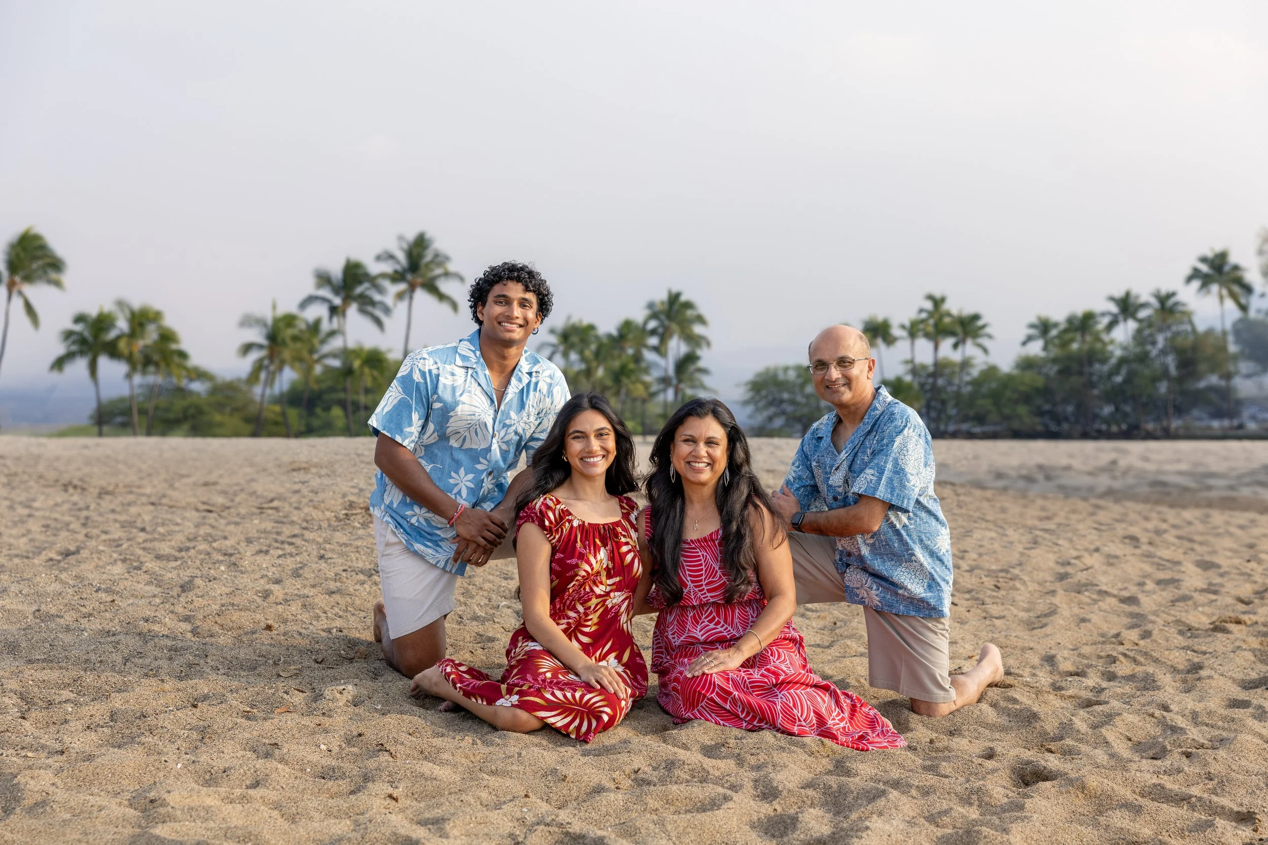 A family of five, including three adults and two children, posing together on a sandy beach with palm trees in the background. They are all smiling and wearing casual, colorful clothing suited for warm weather.