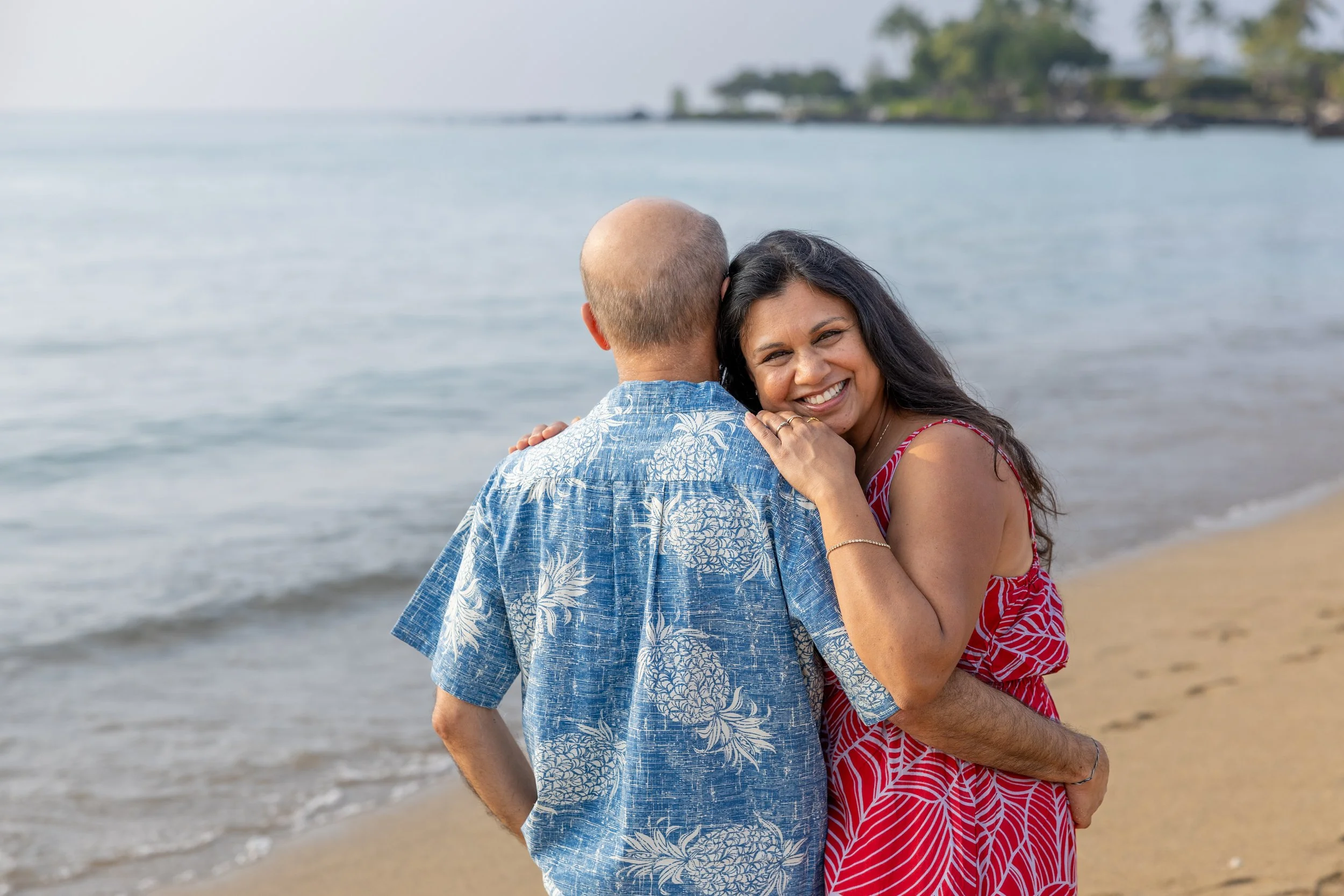 A woman hugging a man at the beach, smiling and looking at the camera, with ocean and distant shoreline in the background.