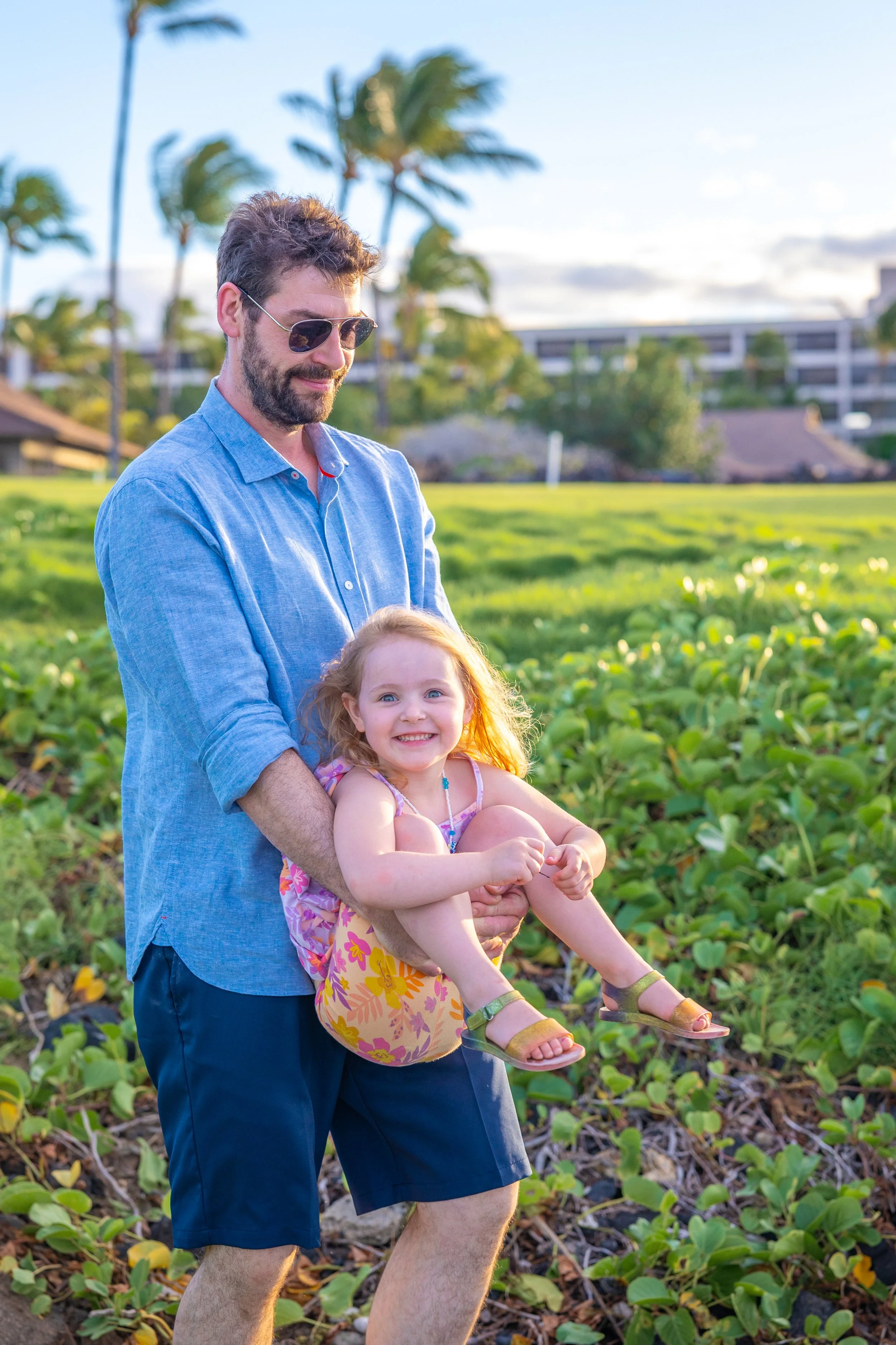 A man in sunglasses and a blue shirt holding a smiling young girl with blonde hair in a floral dress and sandals, standing in a lush green field with palm trees and buildings in the background.