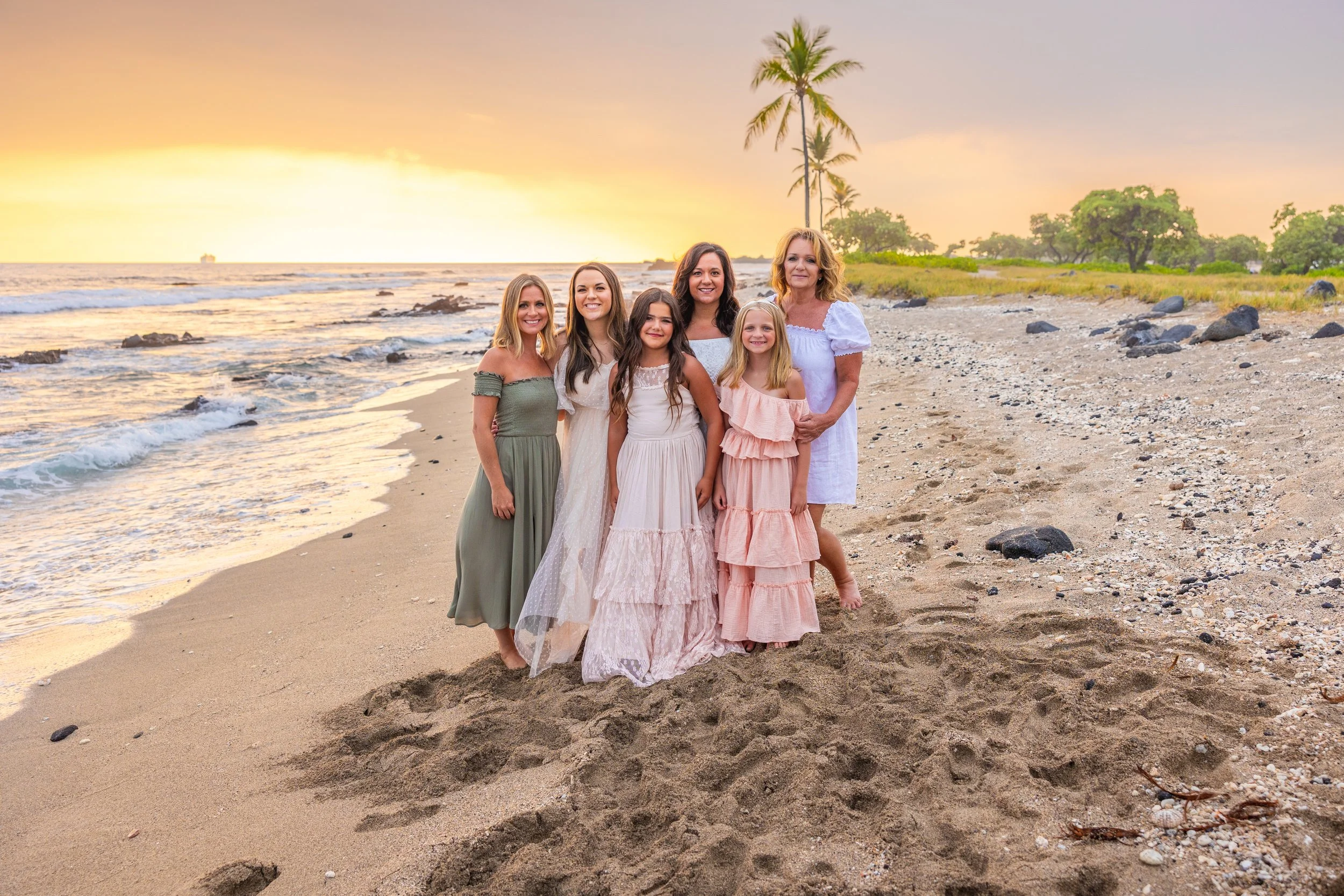 A group of six women and girls standing on a sandy beach at sunset, with palm trees in the background, smiling at the camera.