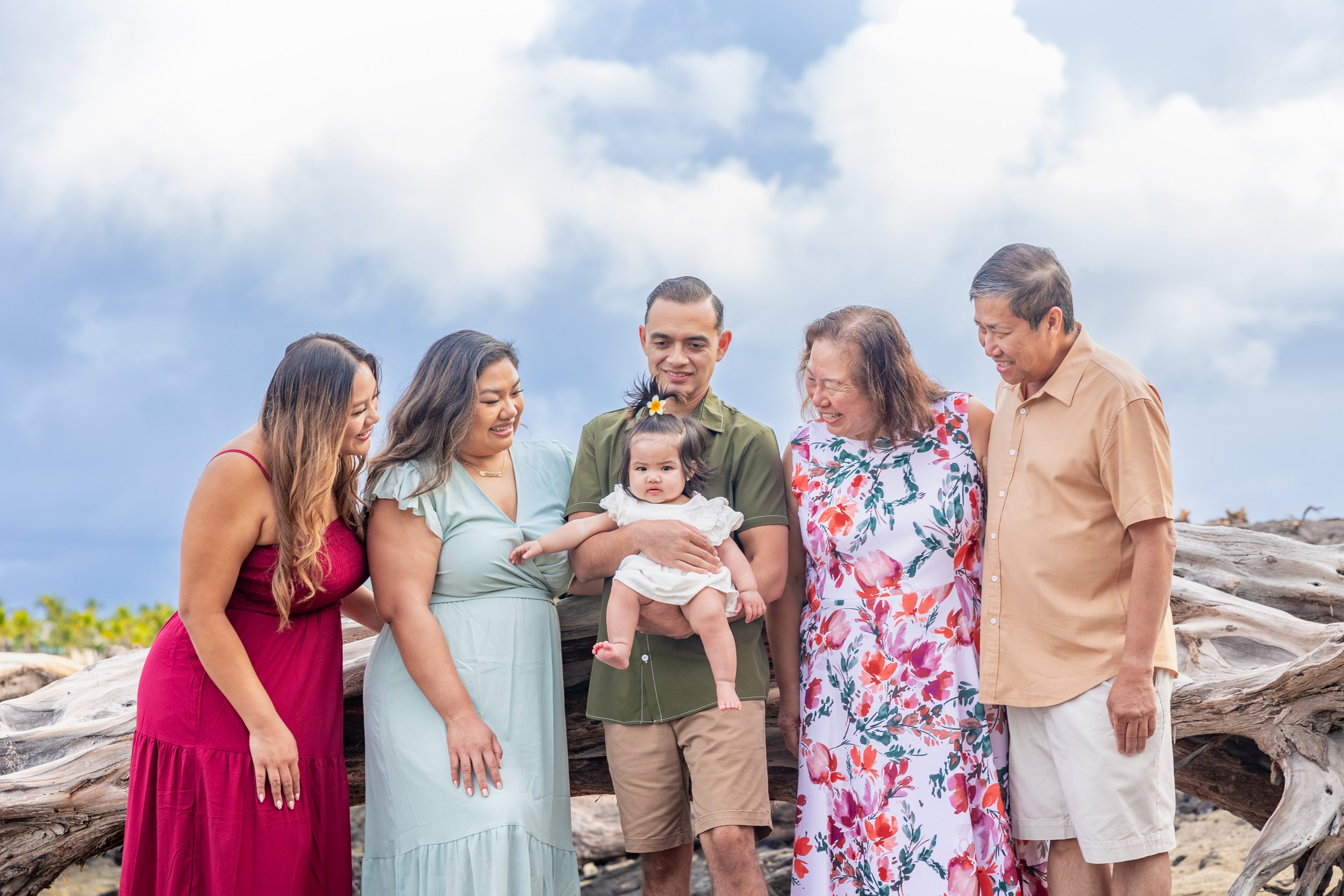 Family group standing outdoors on a beach with driftwood, smiling and looking at a young child being held by a man, dressed in summer clothing, during daytime with cloudy sky.