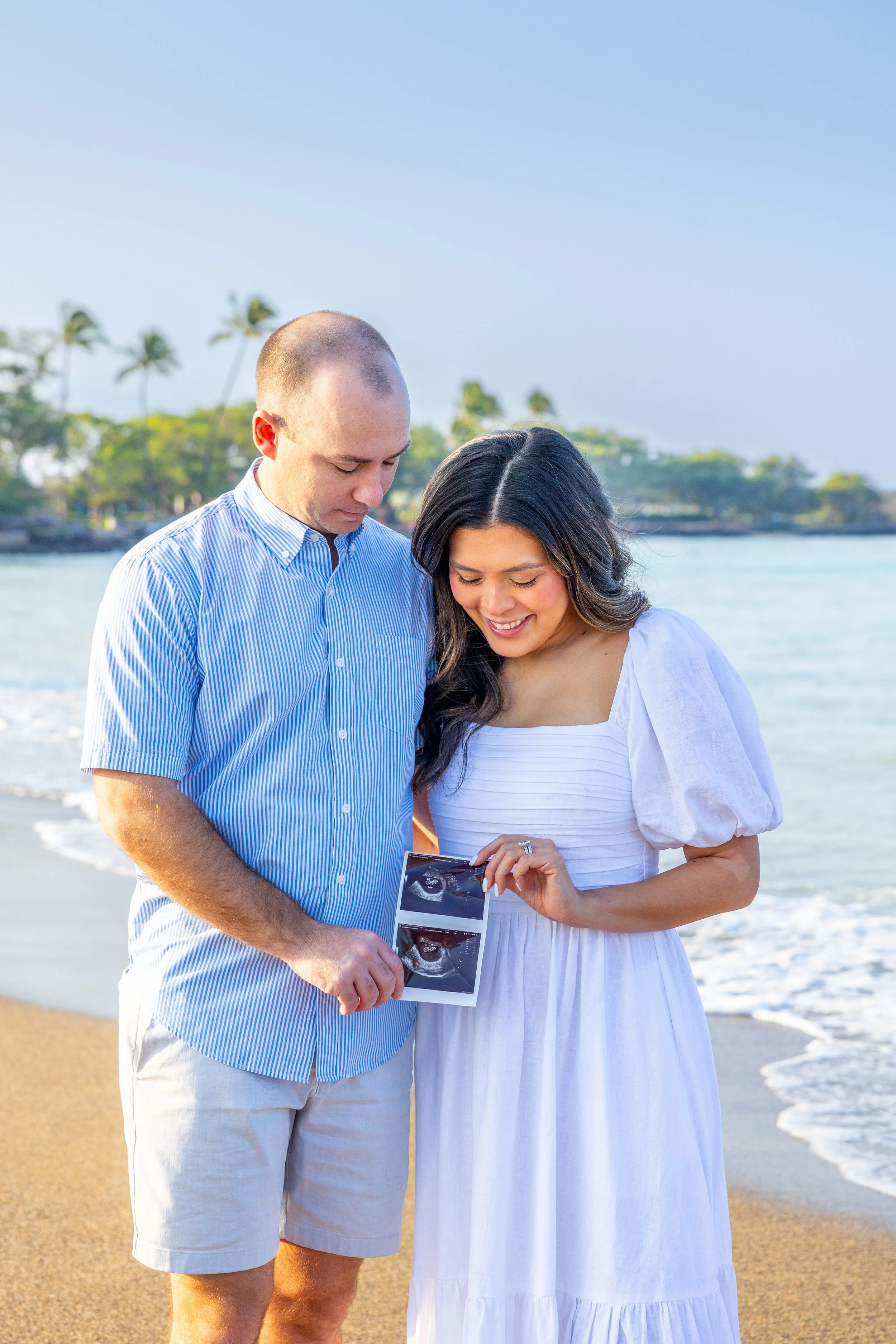Couple on a beach looking at ultrasound photos, woman in white dress, man in blue striped shirt, palm trees and ocean in background.