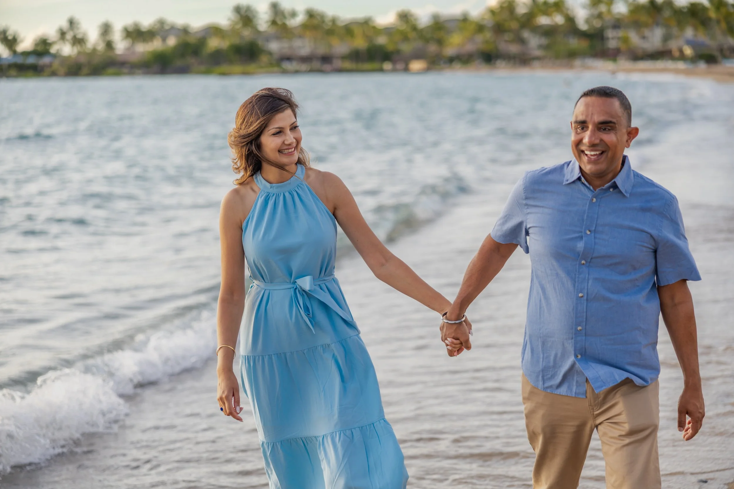 A smiling couple holding hands walking along the beach at sunset, with waves and a distant shoreline in the background.