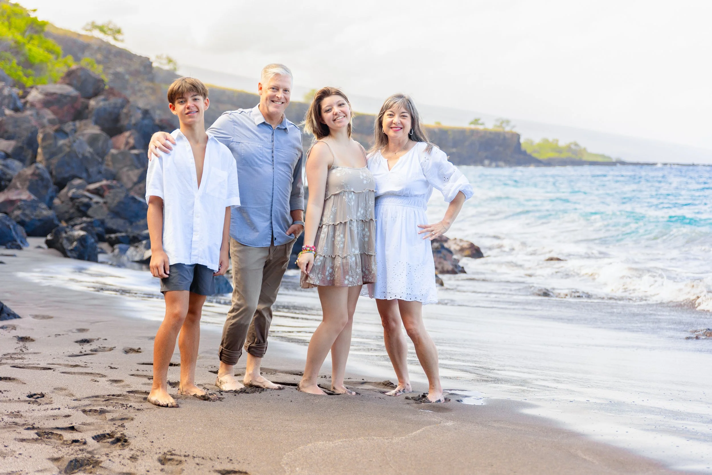 A family of five standing on the beach near the water, smiling and posing for a photo. The scene is bright and sunny with rocks and greenery in the background.