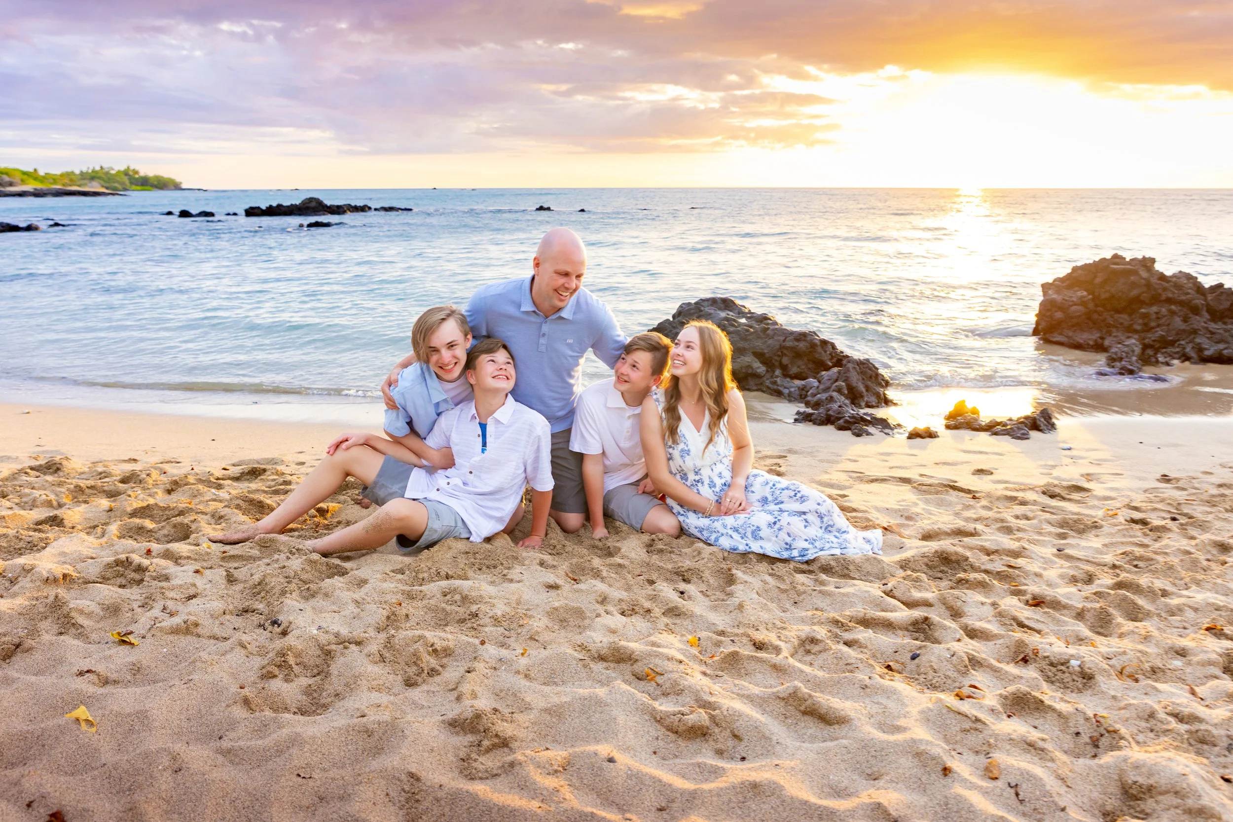 A family of five, including three children and two adults, enjoying a sunset on a sandy beach with rocks and the ocean in the background.