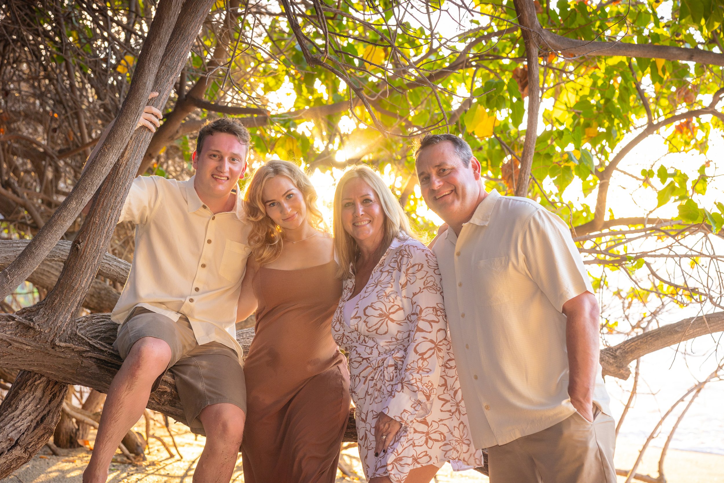 A family of five posing together outdoors under a tree with sunlight filtering through the leaves, on a beach or near water.
