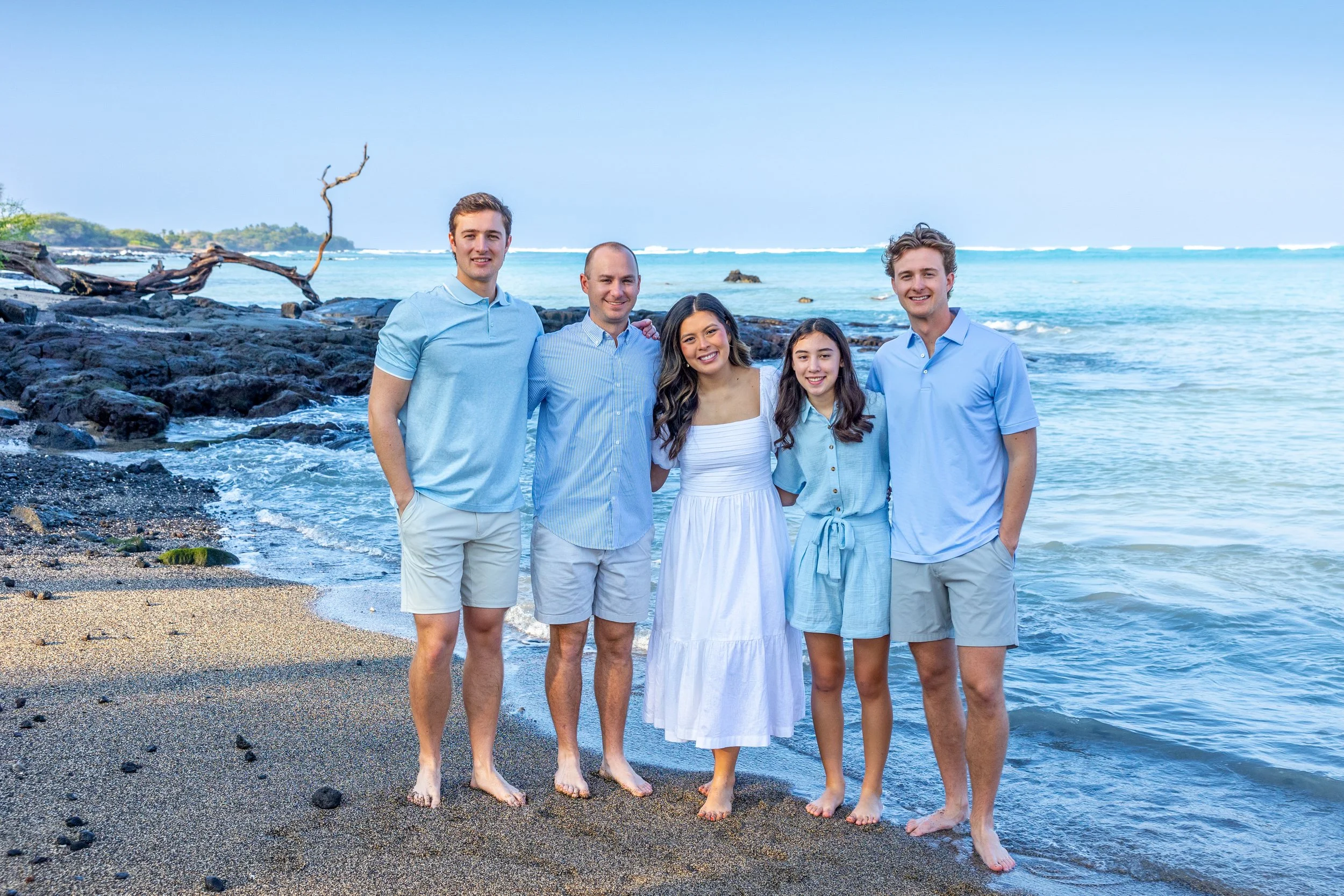 Group of five people standing on a beach near the ocean, smiling and enjoying a sunny day.