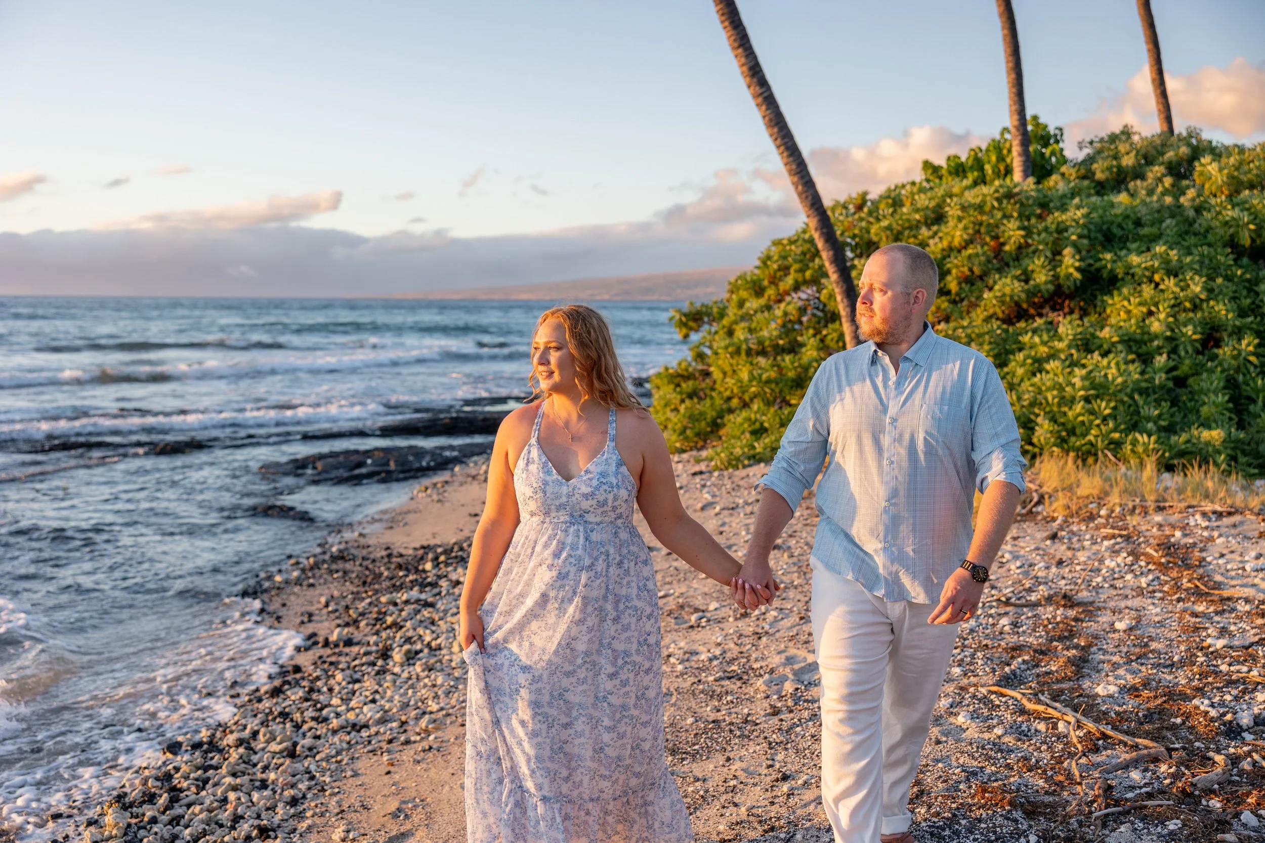 A couple walking hand in hand along a beach during sunset, with palm trees and green bushes in the background.