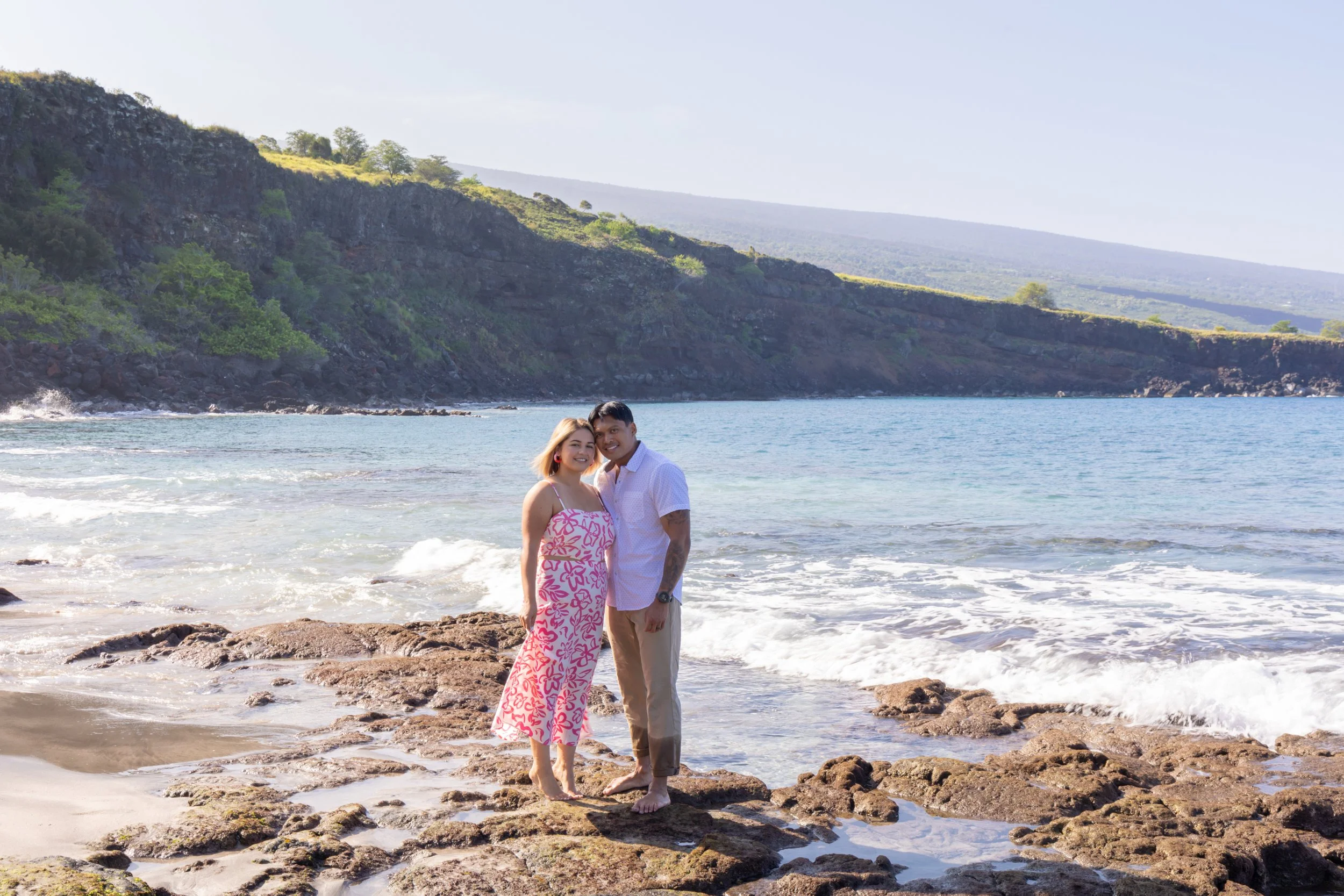 A couple standing on rocks at the beach with waves and green hills in the background.