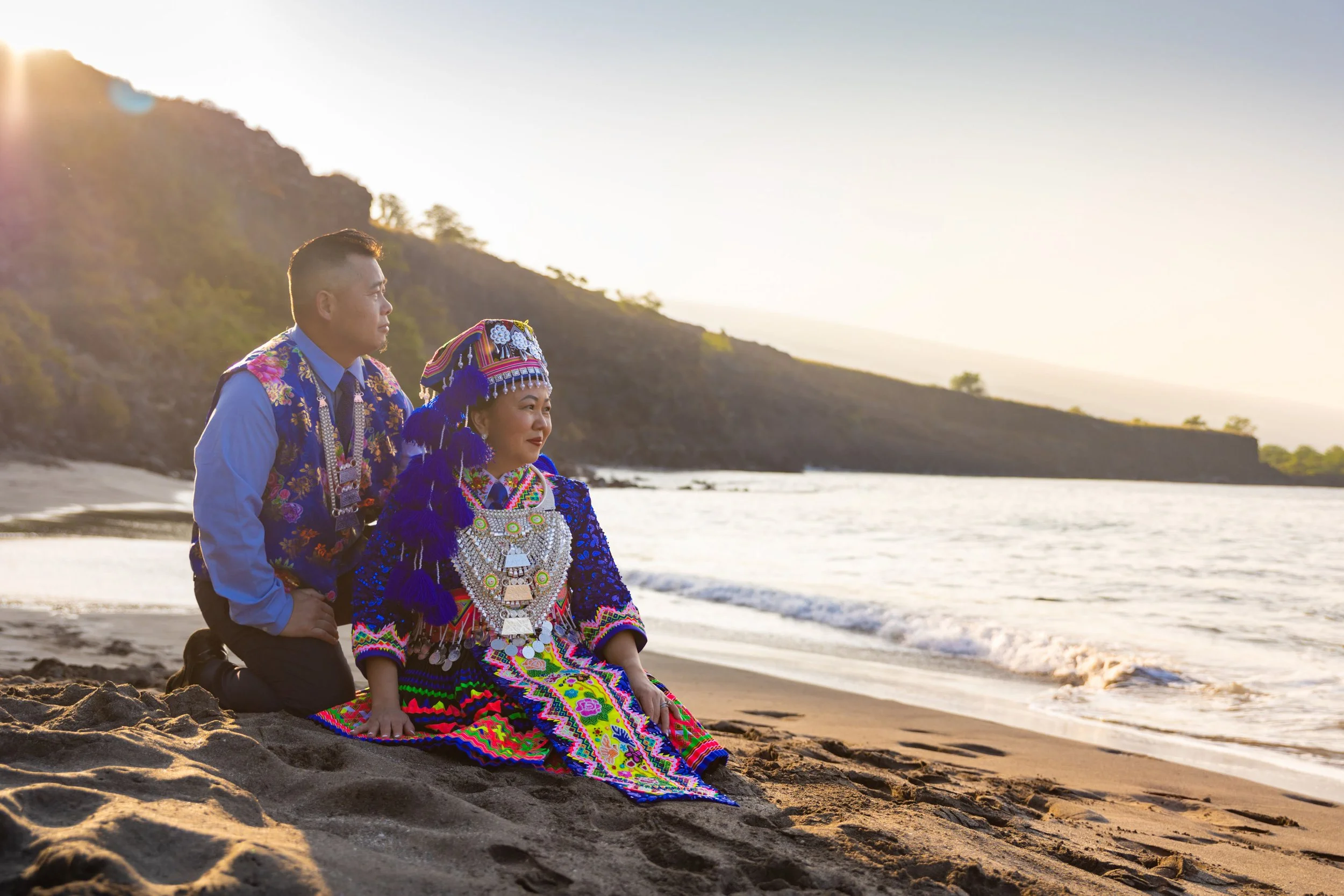 A man and woman in traditional, colorful indigenous clothing kneel on the sand by the ocean at sunset, with hills in the background.
