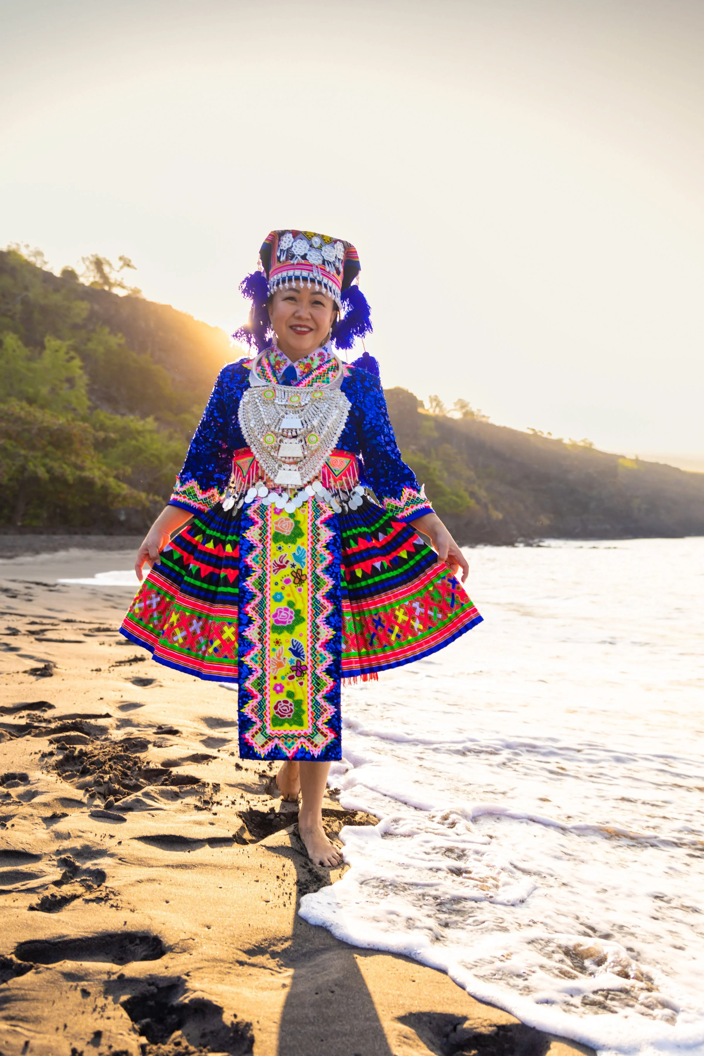 A woman dressed in traditional, colorful attire standing on a beach at sunset with mountains in the background.