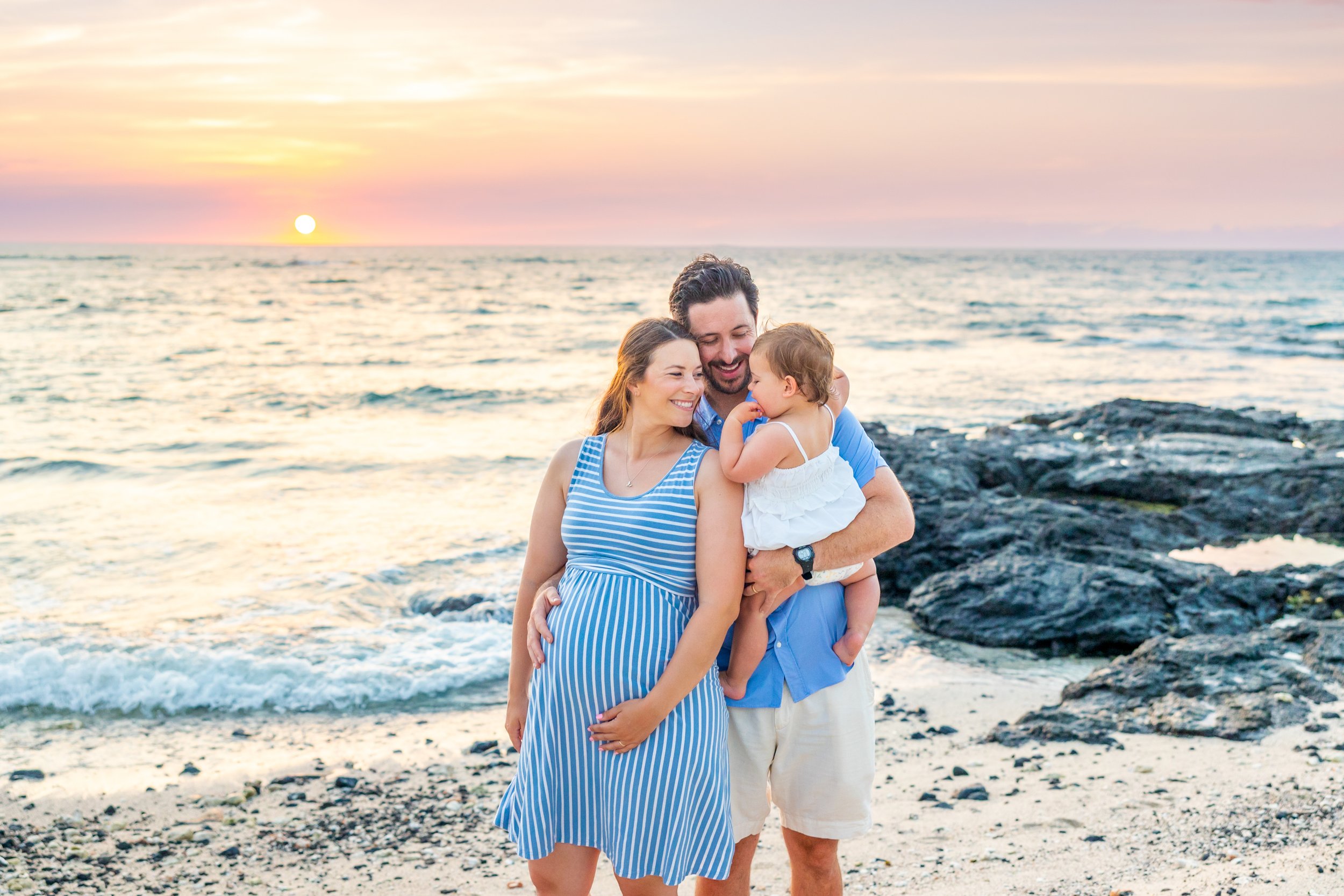 A family of three on a beach during sunset, smiling and embracing each other. The woman is pregnant and wearing a blue and white striped dress, the man is wearing a blue shirt and white shorts, holding a young girl in a white dress. The background sh