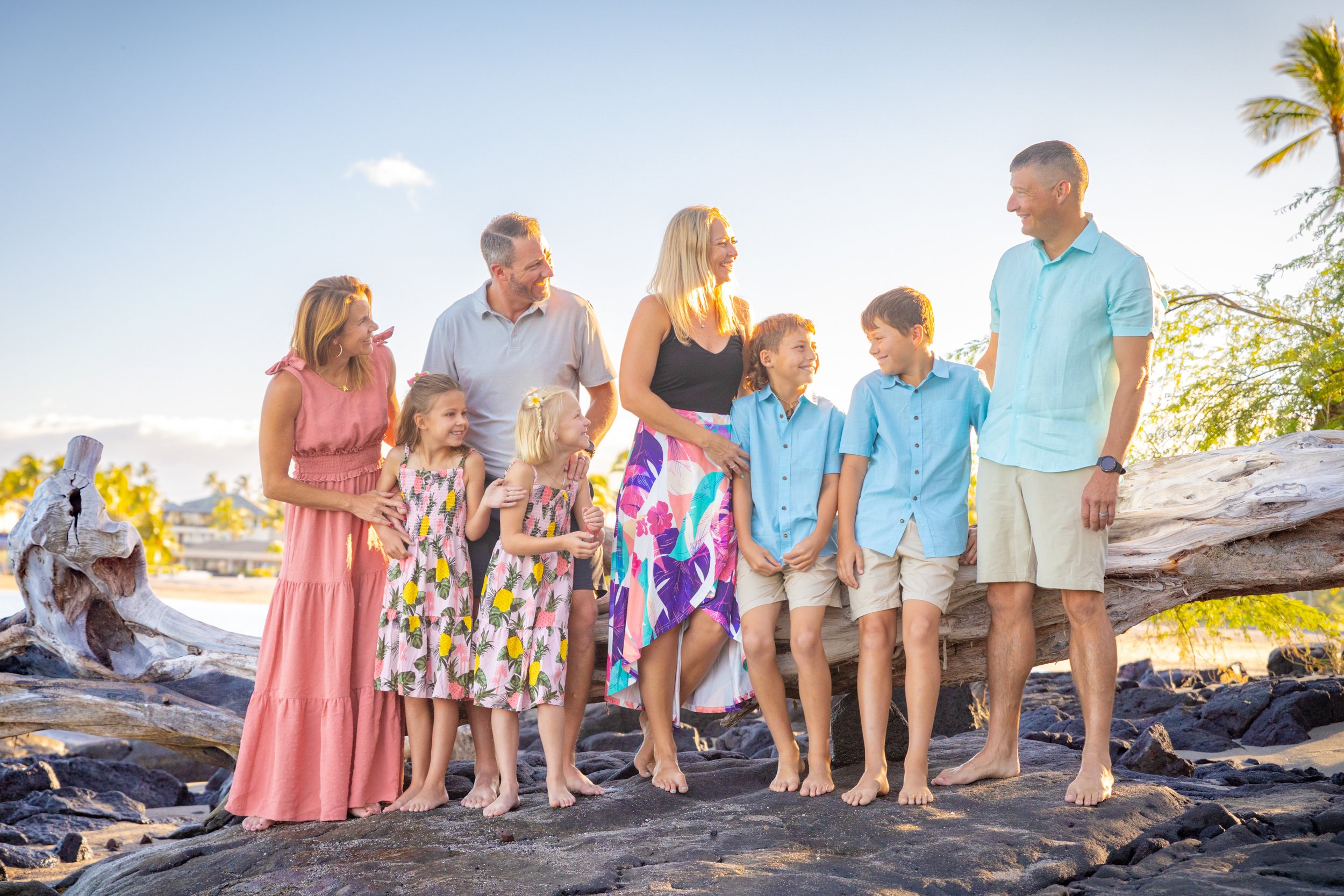 A family of ten, including three adults and seven children, standing on a rocky beach with driftwood, palm trees, and a sunset sky in the background, smiling and enjoying time together.
