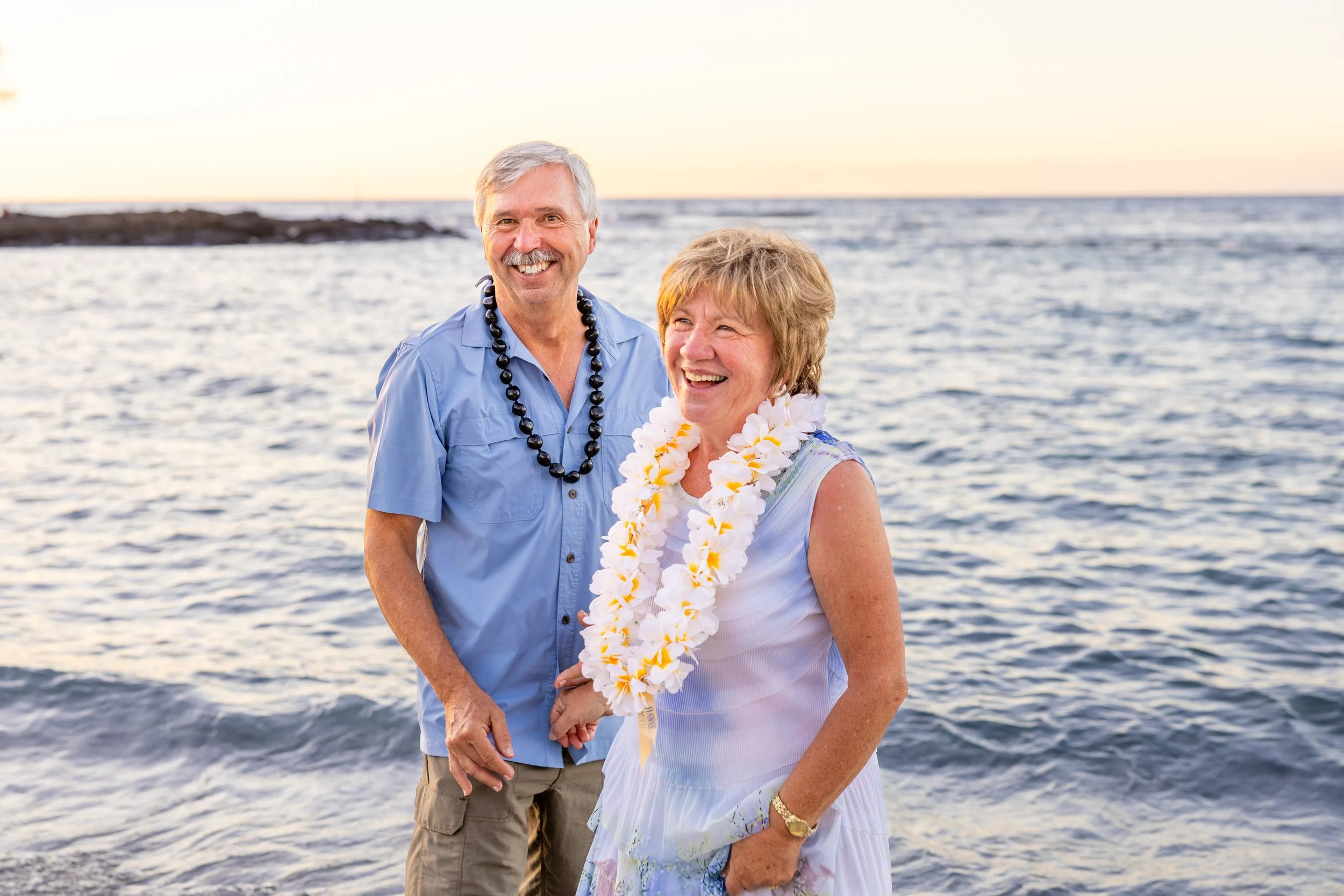 An elderly couple smiling at the beach during sunset, holding hands, wearing leis and casual summer clothing.