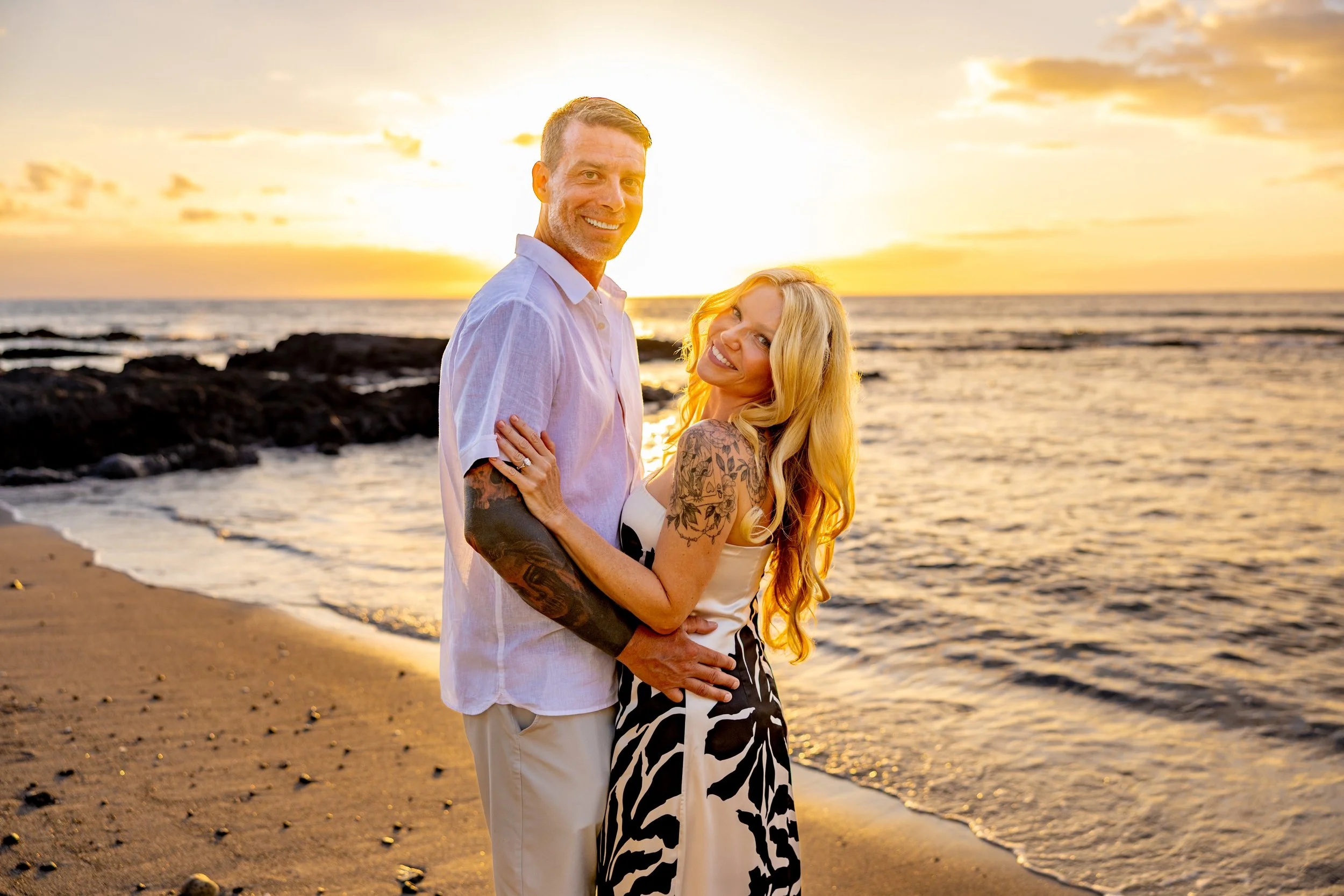 A happy couple standing on the beach during sunset, smiling and embracing each other, with the ocean and rocky shoreline in the background.