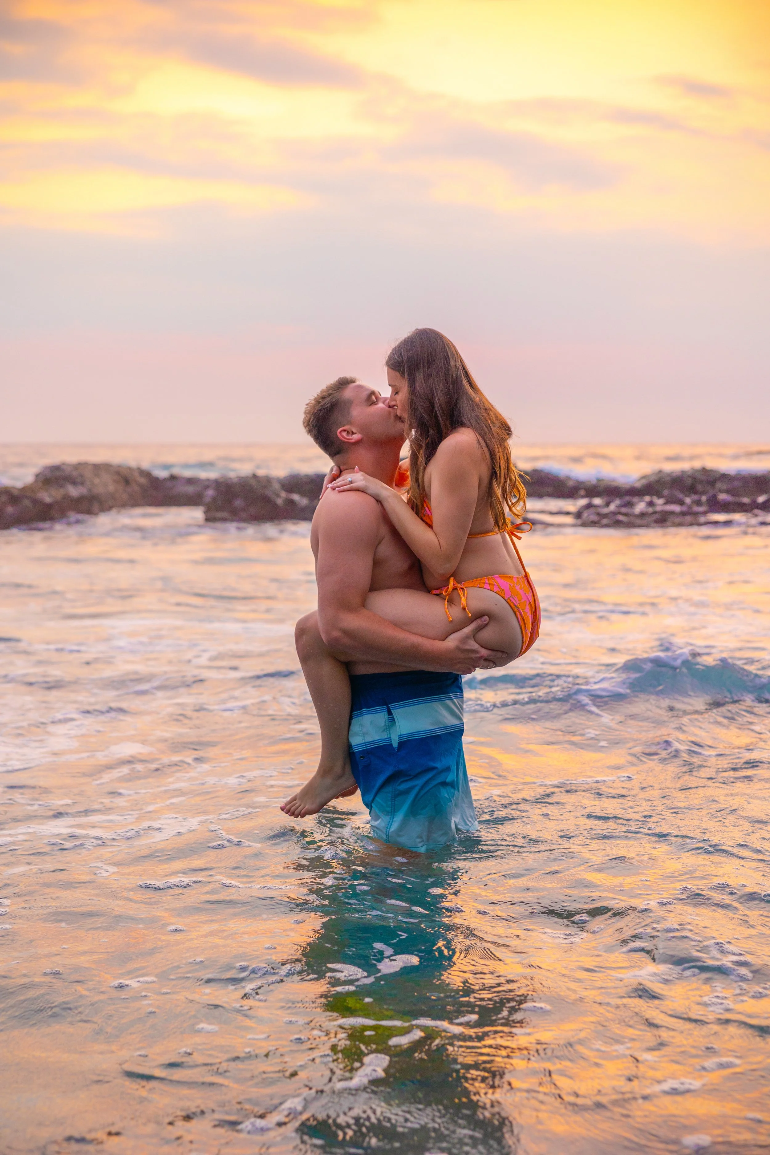 A couple kissing in the ocean at sunset, with the man holding the woman in his arms, both wearing swimwear.
