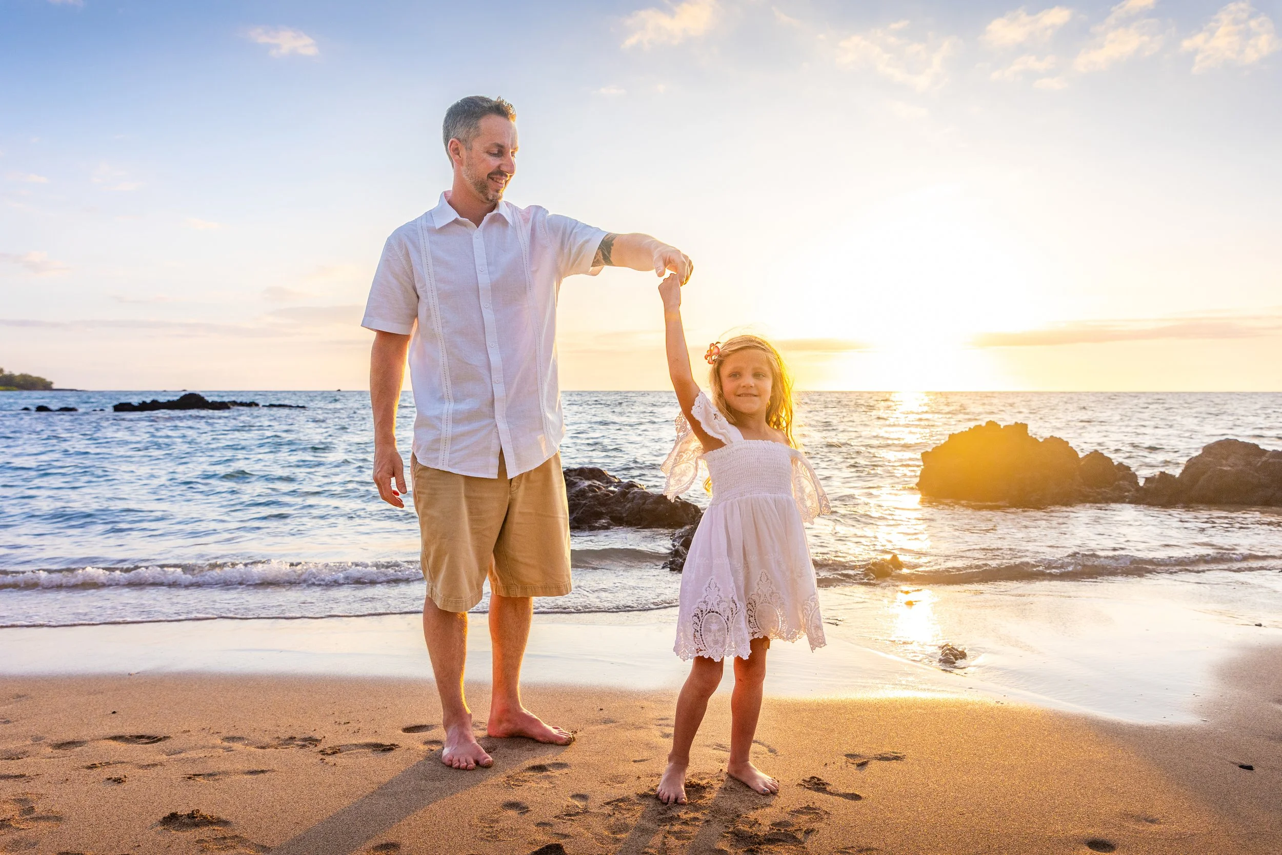 A man and young girl holding hands on a beach at sunset, with the man lifting the girl's hand in the air. The girl is smiling and wearing a white dress. The scene features ocean waves, rocks, and a bright sky with clouds in the background.