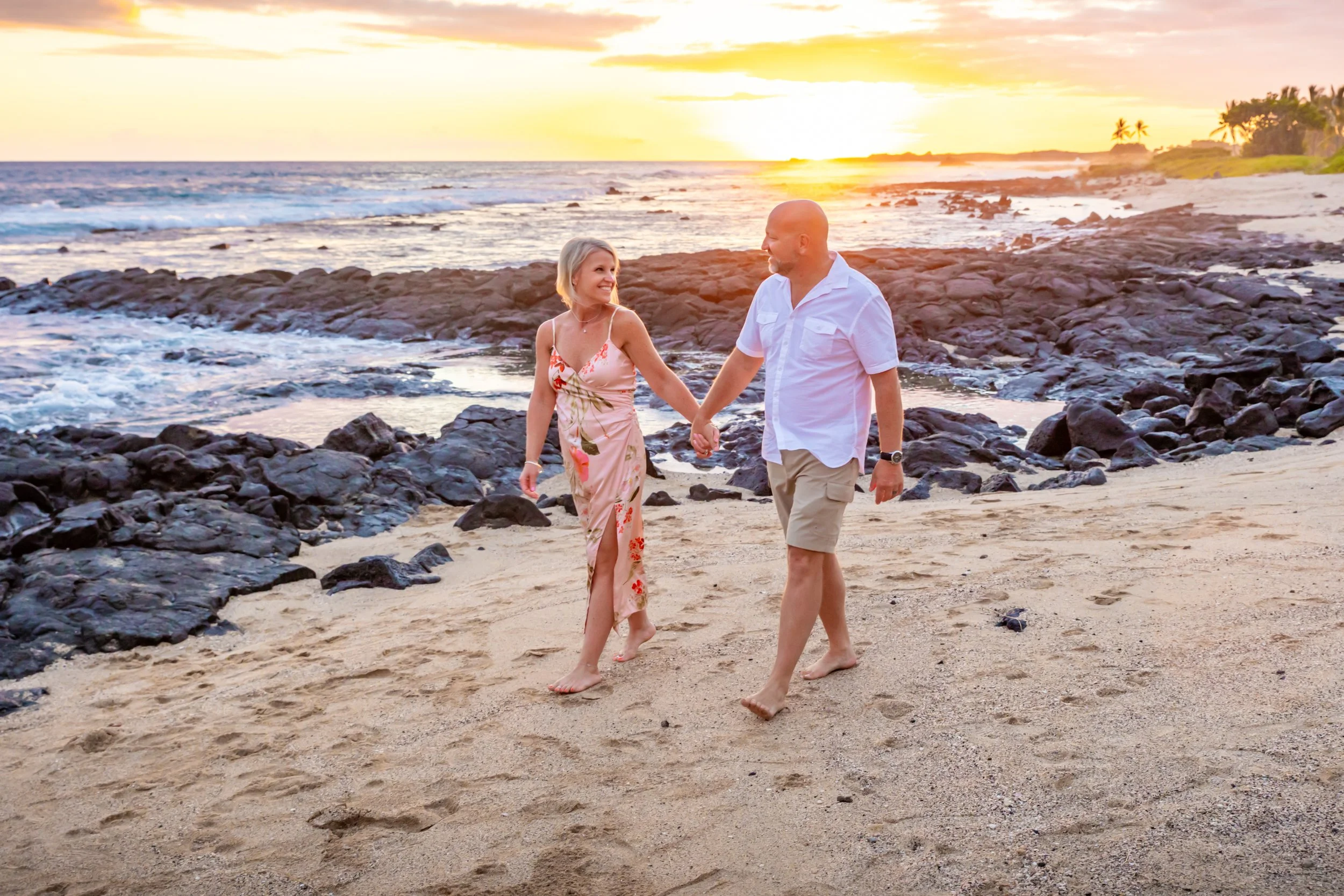 A couple walking hand in hand on a beach at sunset, with rocks, ocean waves, and palm trees in the background.