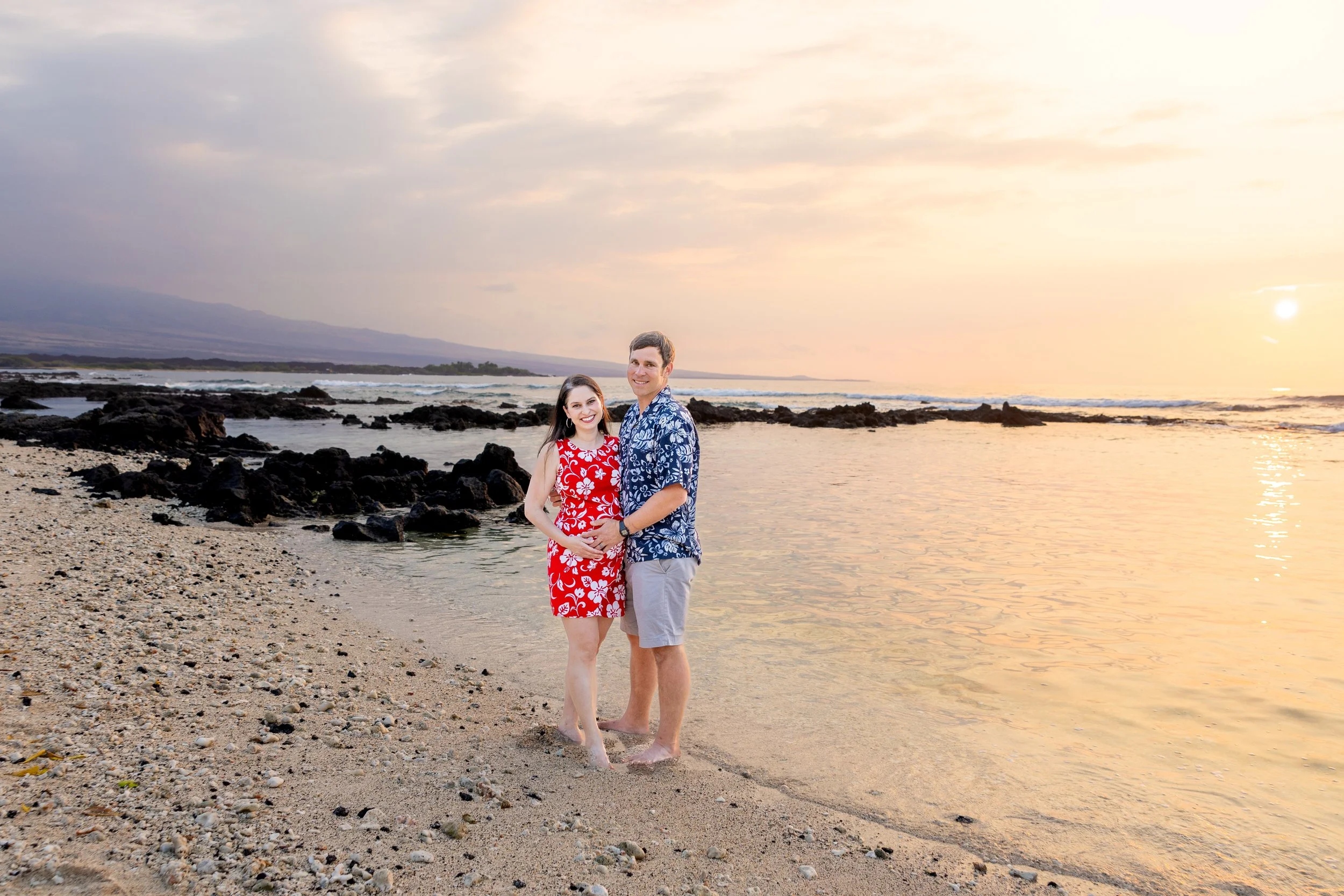 A smiling couple stands on a beach at sunset, holding hands, with rocky shoreline and calm ocean waves in the background.