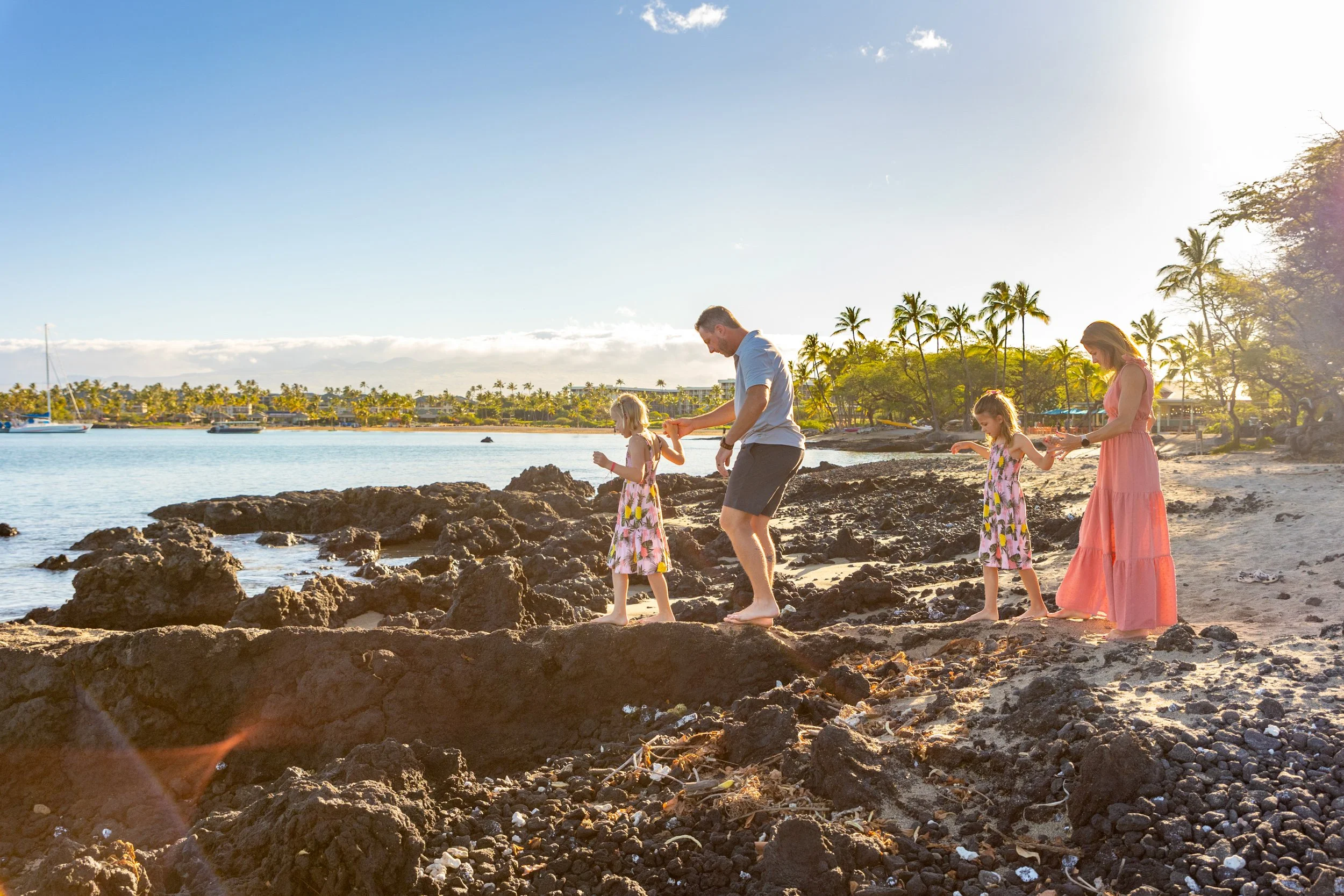 Family of four, including a man, a woman, and two young girls, exploring rocky coastline at sunset, with sailboats on the water and palm trees in the background.