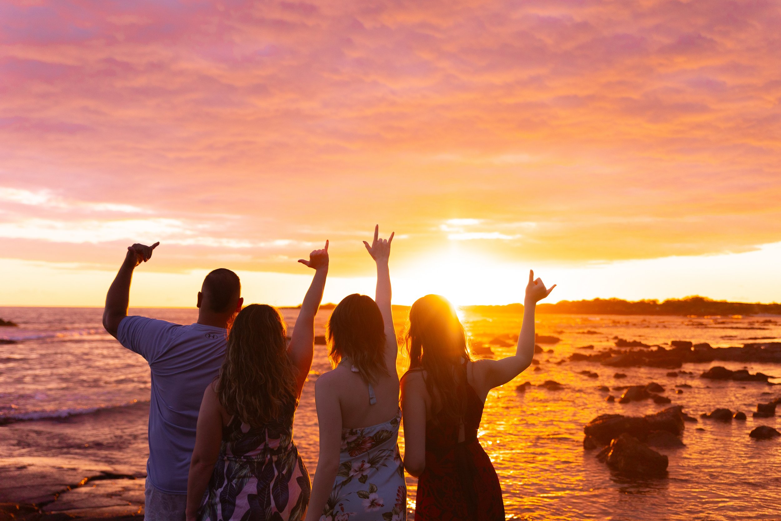 Four people standing on a beach at sunset, silhouetted against the colorful sky, with one person making a rock on hand gesture and another pointing upward.