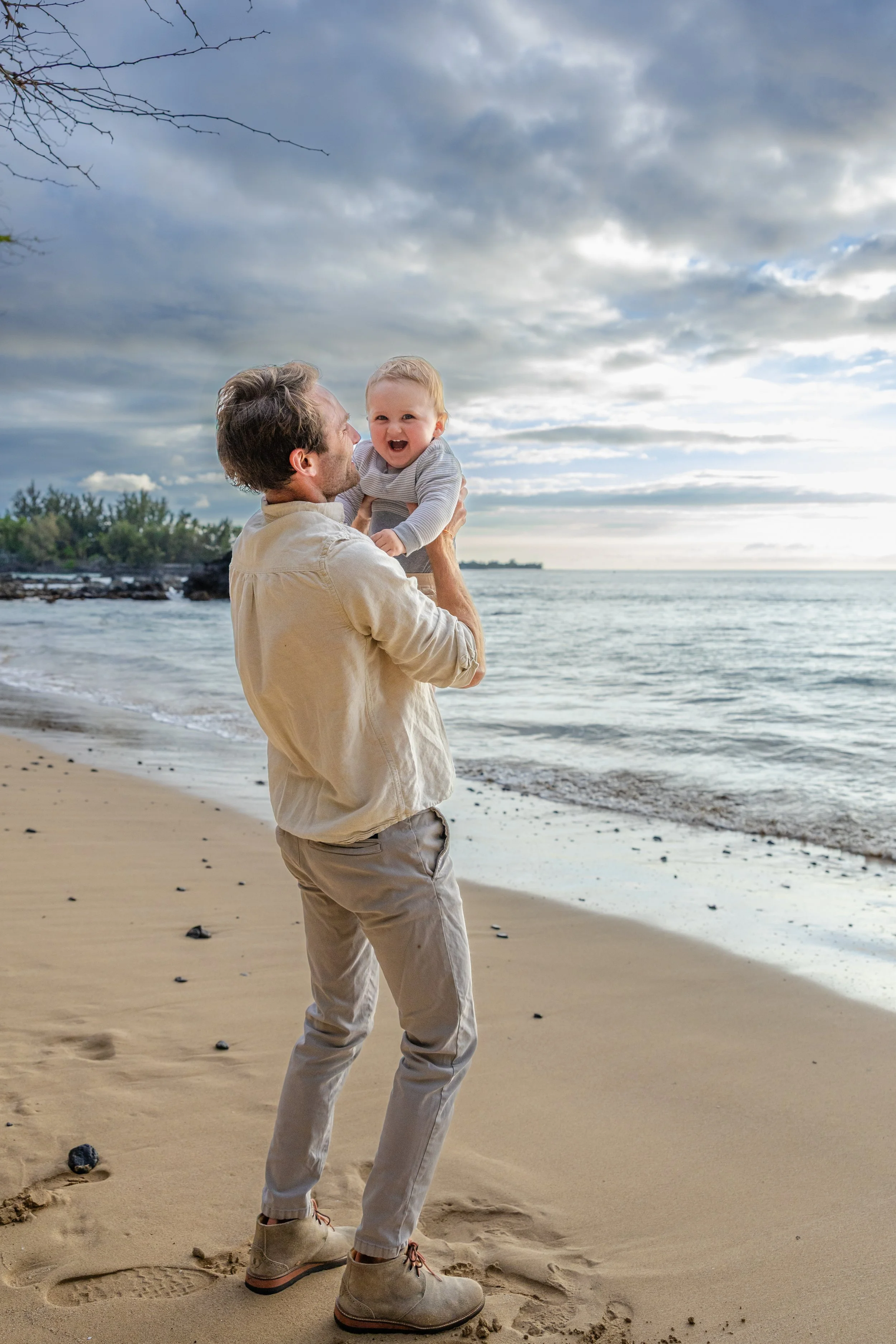 A man holding a smiling toddler girl on a beach with cloudy sky and ocean in the background.