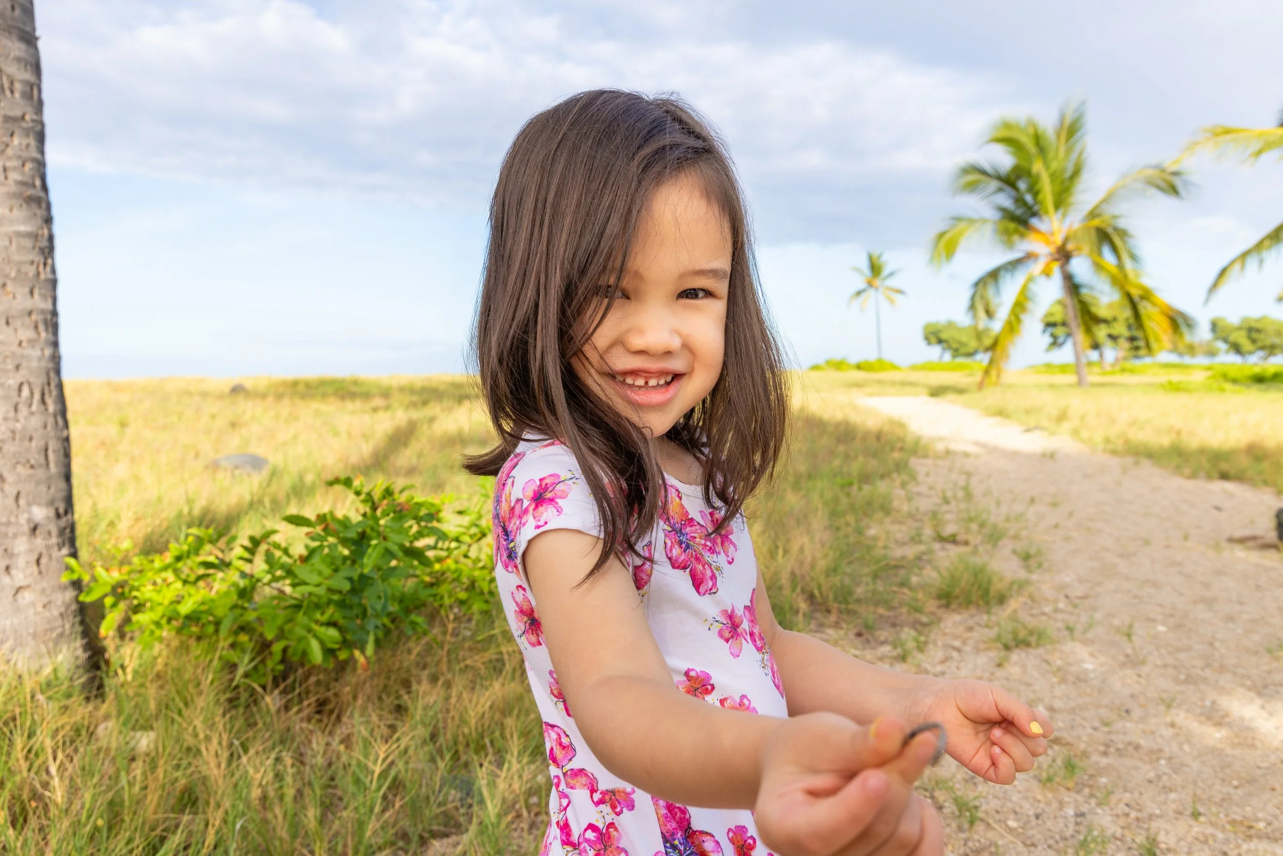 Smiling young girl in a floral dress holding a small object in an outdoor tropical setting with palm trees and grassy trail.