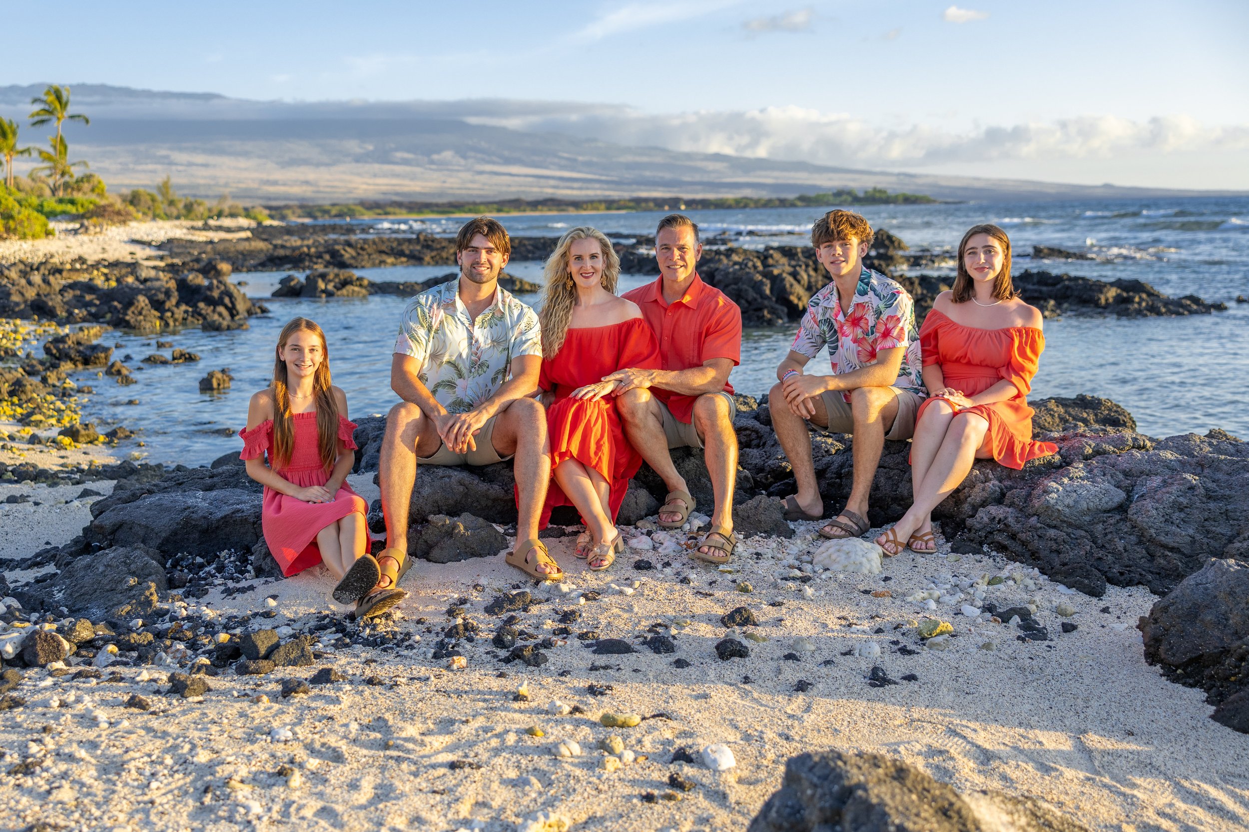 A family of seven sitting on rocks at the beach with the ocean in the background, wearing colorful summer clothes.