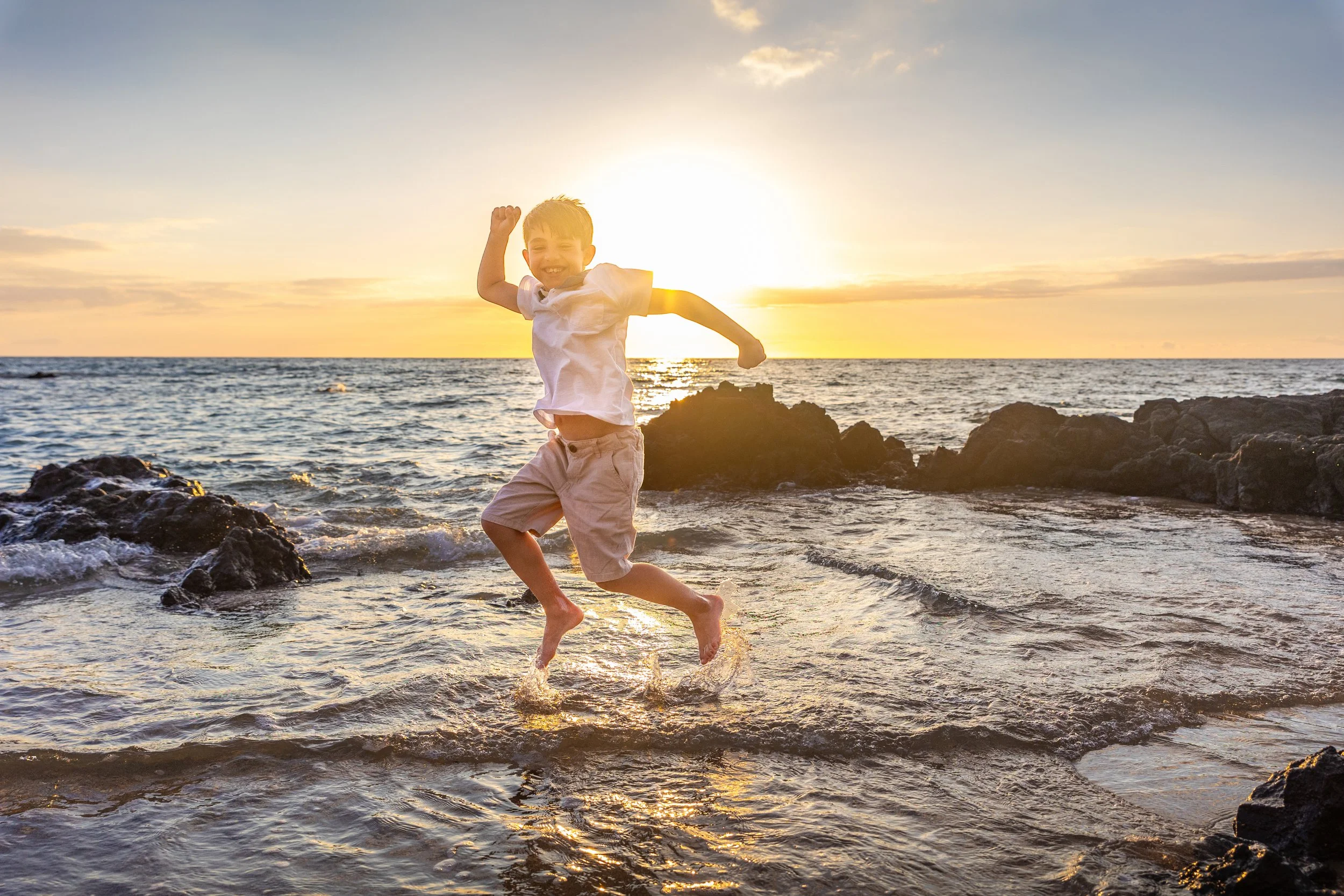 A smiling young boy jumping in the shallow waters of a beach at sunset, with rocks and the ocean in the background.