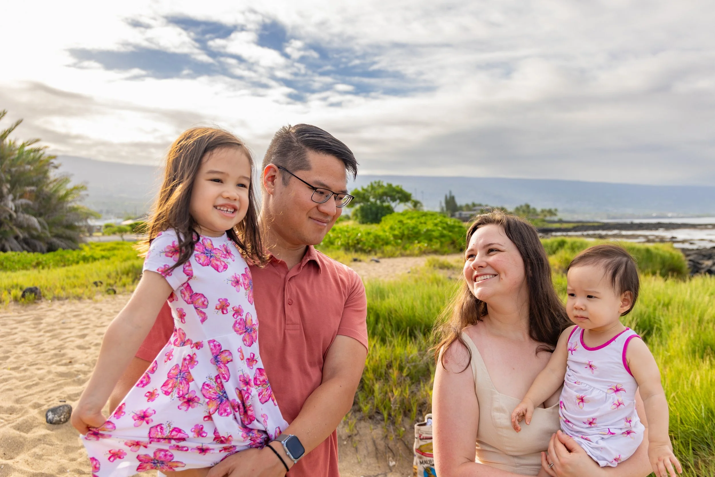 A family of four enjoying a day at the beach, with a man holding a young girl and a woman holding a toddler girl, all smiling and standing on sandy and grassy terrain with a cloudy sky and distant mountains in the background.