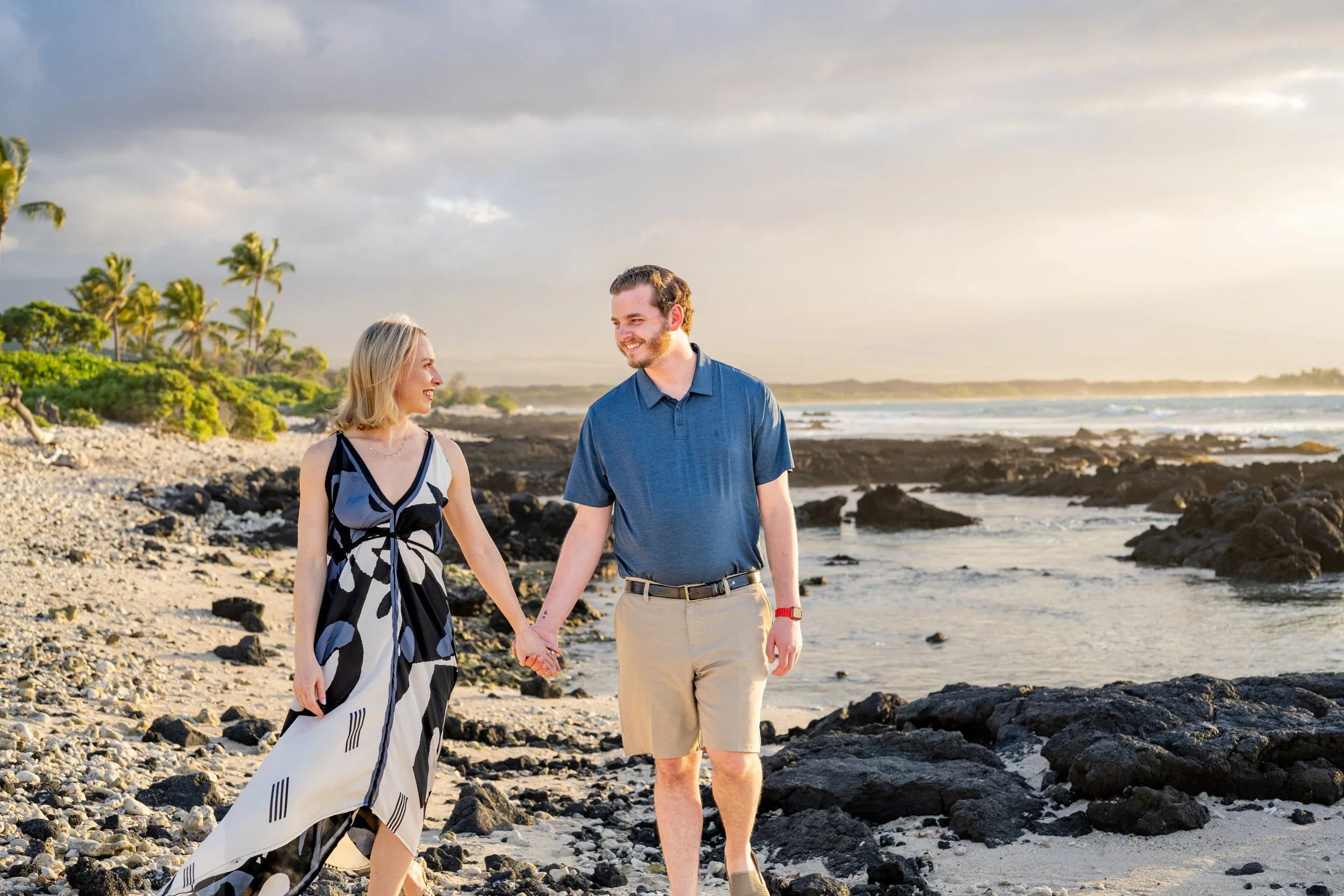 A young couple holding hands and walking along a rocky beach during sunset, with palm trees and ocean waves in the background.